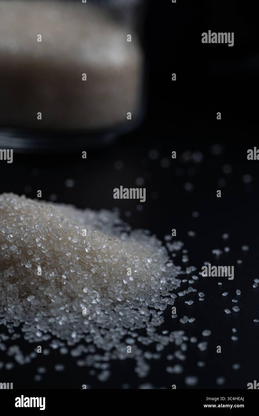 Sucre granulé blanc renversant d'un bocal en verre sur une surface sombre. Parfait pour la photographie alimentaire, la conception d'emballage ou le contenu lié à la cuisine Banque D'Images
