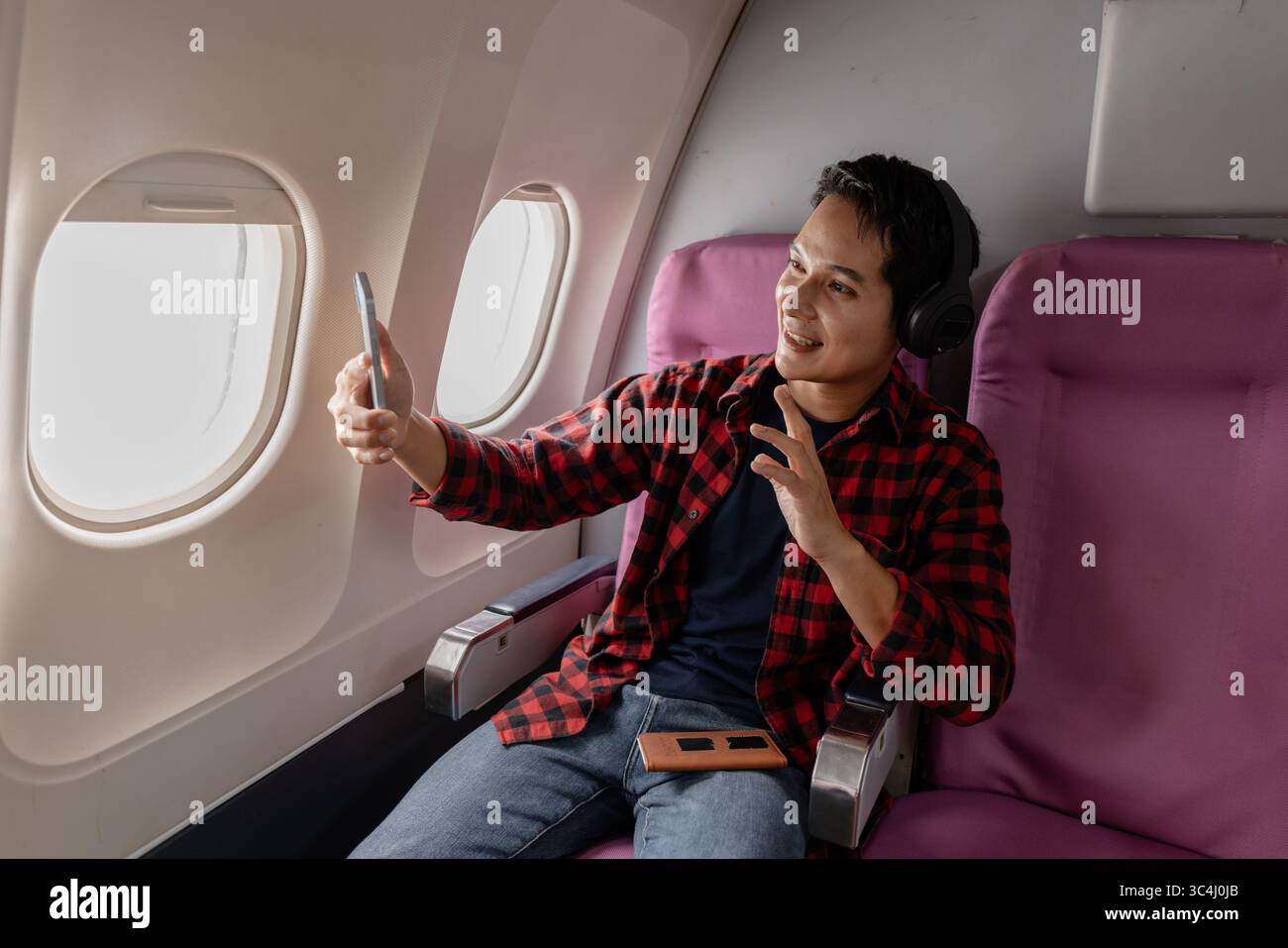 Un jeune homme joyeux aime son vol tout en utilisant son smartphone et en portant des écouteurs. Assis près de la fenêtre de l'avion, il montre de l'excitation et happ Banque D'Images