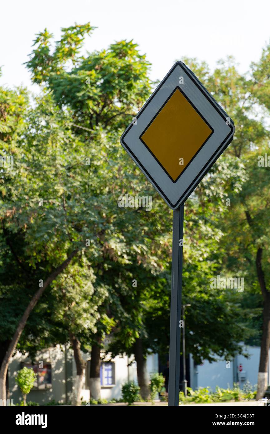 Un panneau routier prioritaire en forme de losange jaune avec une bordure blanche, posé contre des arbres verdoyants et un bâtiment dans un environnement urbain Banque D'Images