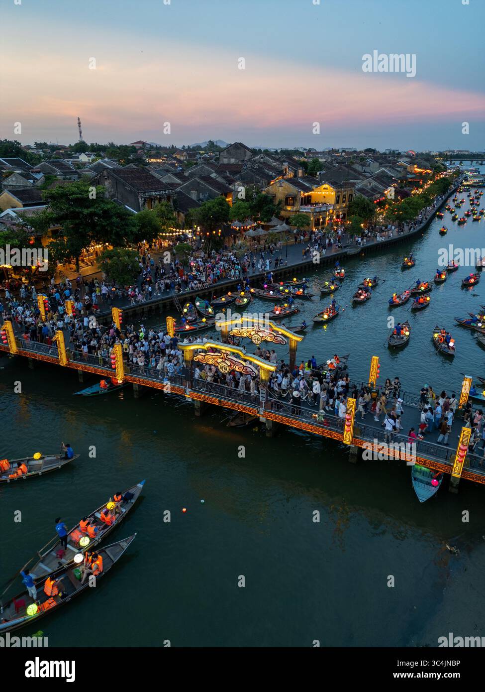 Vue aérienne d'un pont éclairé par des lumières chaudes, animé par des gens et des bateaux dérivant le long de la rivière, reflétant le ciel crépusculaire, Hội an, Quảng Nam, Vietnam. Banque D'Images