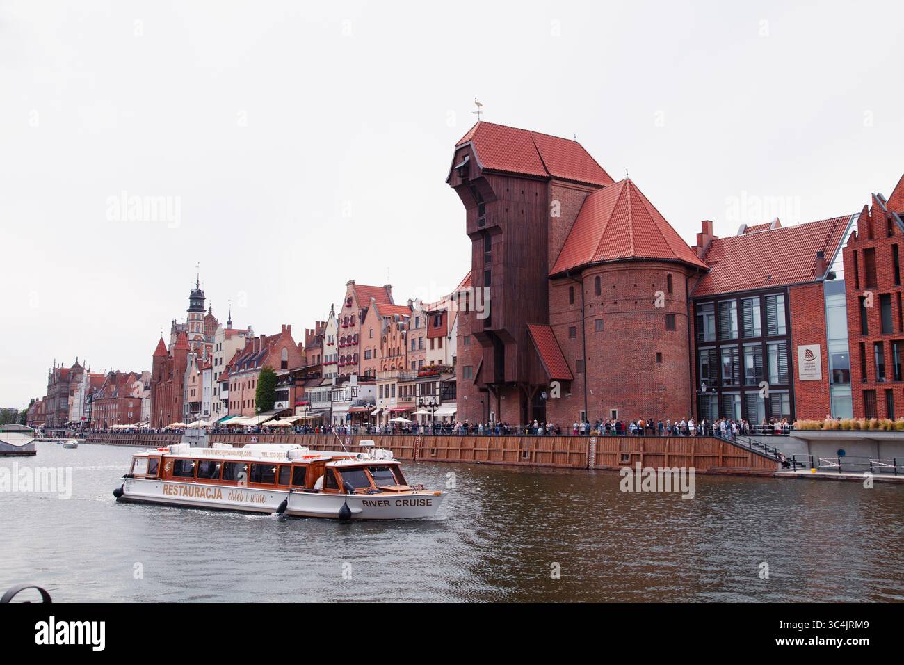 Gdansk, Pologne - 04-07-2025 - Un bateau-restaurant naviguant le long du canal avec la grue historique de la porte de la ville en arrière-plan Banque D'Images