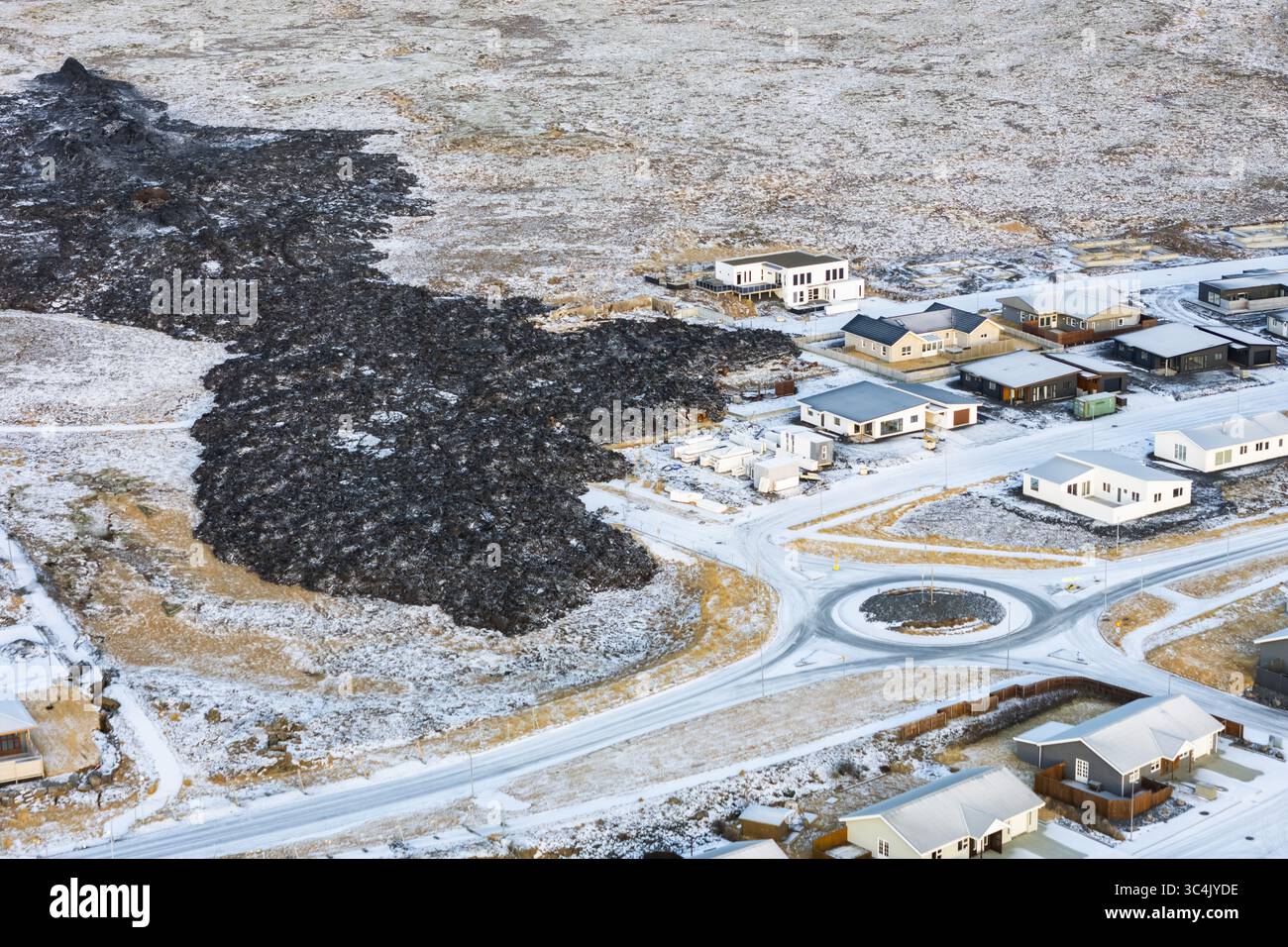 Vue aérienne d'une coulée de lave durcie noire et dure empiétant sur une ville poussiéreuse de neige, un contraste spectaculaire de destruction et de résilience, Grindavik, Grindavíkurbær, Islande. Banque D'Images