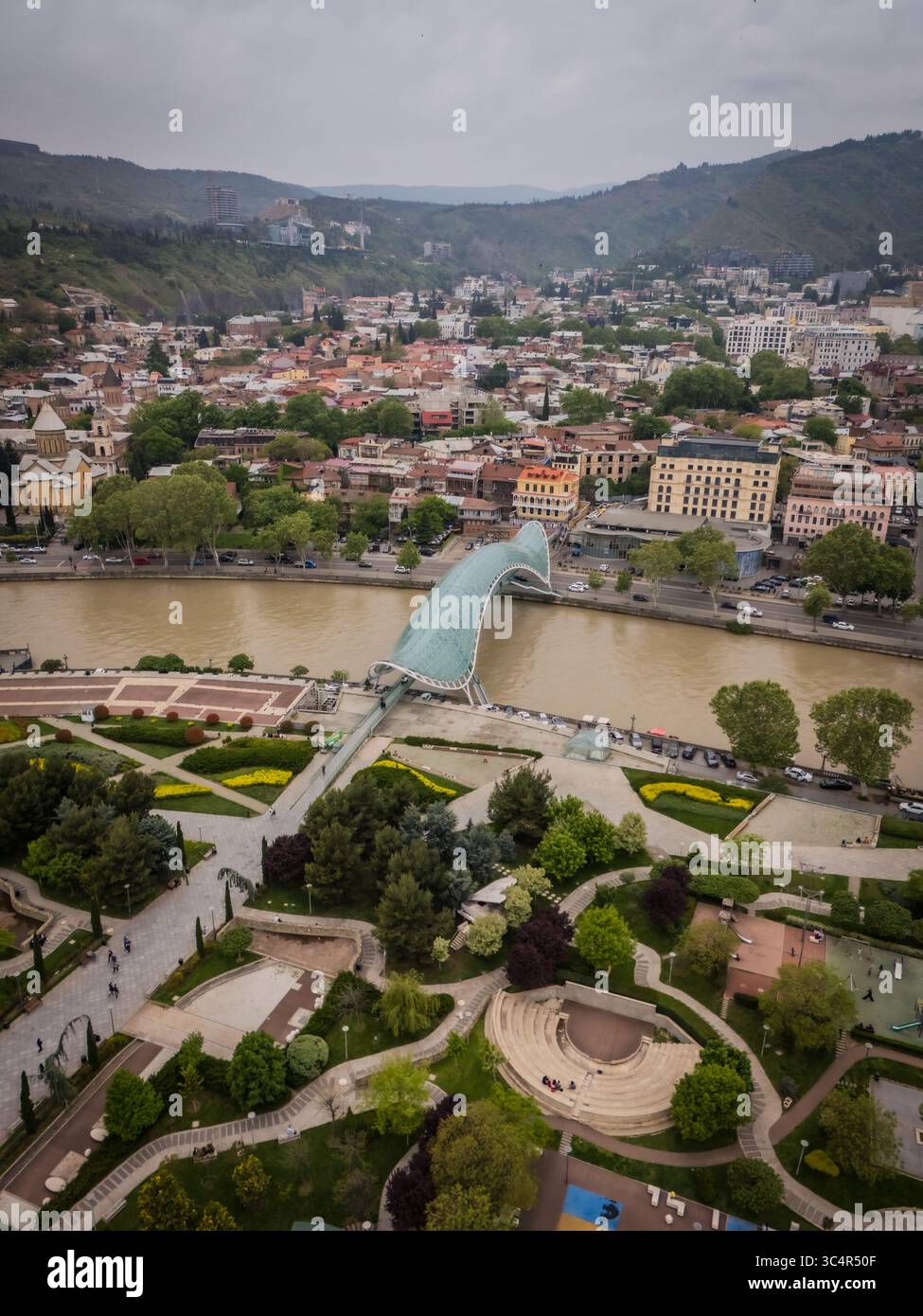 Vue aérienne du Pont de la paix brillante sur la rivière Kura boueuse, embrassée par des parcs verdoyants et l'étendue urbaine nichée dans la vallée, Tbilissi, Tbilissi, Géorgie. Banque D'Images