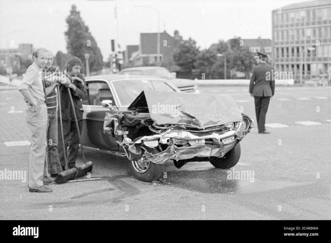 Coin accident Randweg - Leidsevaart, accidents, collisions, accidents, 04-09-1975 Whizgle News, Dutch Desk, pays-Bas, 1950-2000 Une voiture lourdement endommagée se trouve à une intersection, avec son avant froissé et des débris dispersés sur la route. Deux individus, dont l'un tient un balai, évaluent les lieux, tandis qu'un policier se tient à proximité pour observer la situation. Les bâtiments environnants peuvent être vus en arrière-plan, avec le trafic au loin. Banque D'Images