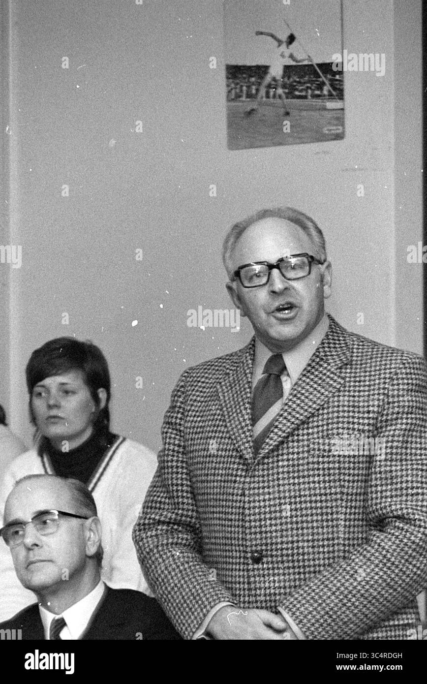 Actions chez CIO entraînement sportif chez Duinlust Whizgle News, Dutch Desk, pays-Bas, 1950-2000 Un homme portant des lunettes et un costume à motifs se tient devant, parlant passionnément, tandis que deux personnes derrière lui écoutent attentivement. L'une est une femme aux cheveux courts, et l'autre est un homme plus âgé en costume. Une affiche sportive est accrochée au mur en arrière-plan. Banque D'Images