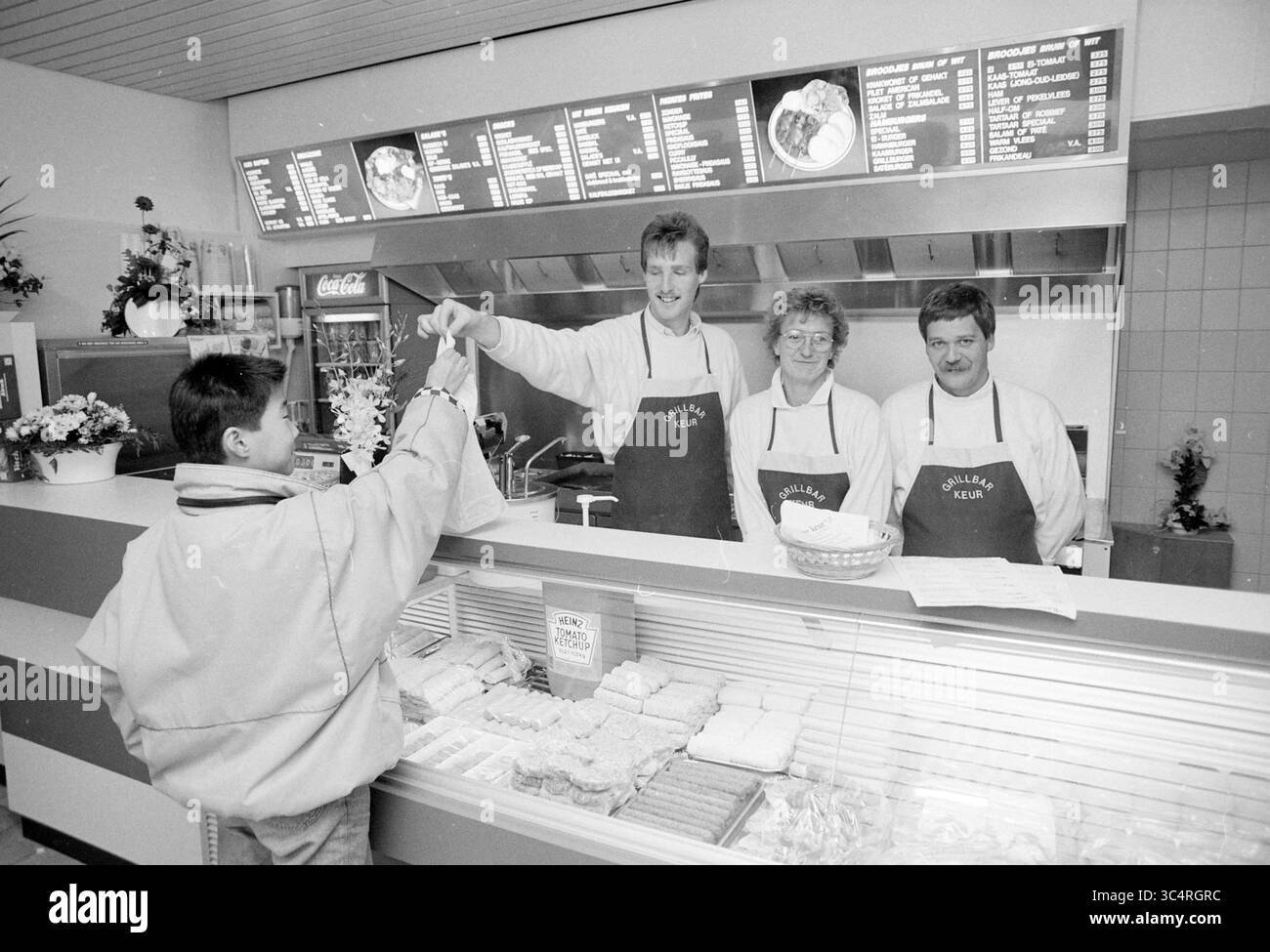 Grill bar Keur Heemstede, intérieur avec trois employés, Heemstede, pays-Bas, 21-11-1990 Whizgle News, Dutch Desk, pays-Bas, 1950-2000 Une équipe joyeuse de trois membres du personnel se tient derrière un comptoir dans un établissement alimentaire, souriant alors qu'ils servent un client qui tend la main pour sa commande. Le menu est affiché ci-dessus, présentant divers produits alimentaires, tandis que le comptoir est bordé de produits fraîchement préparés. Banque D'Images