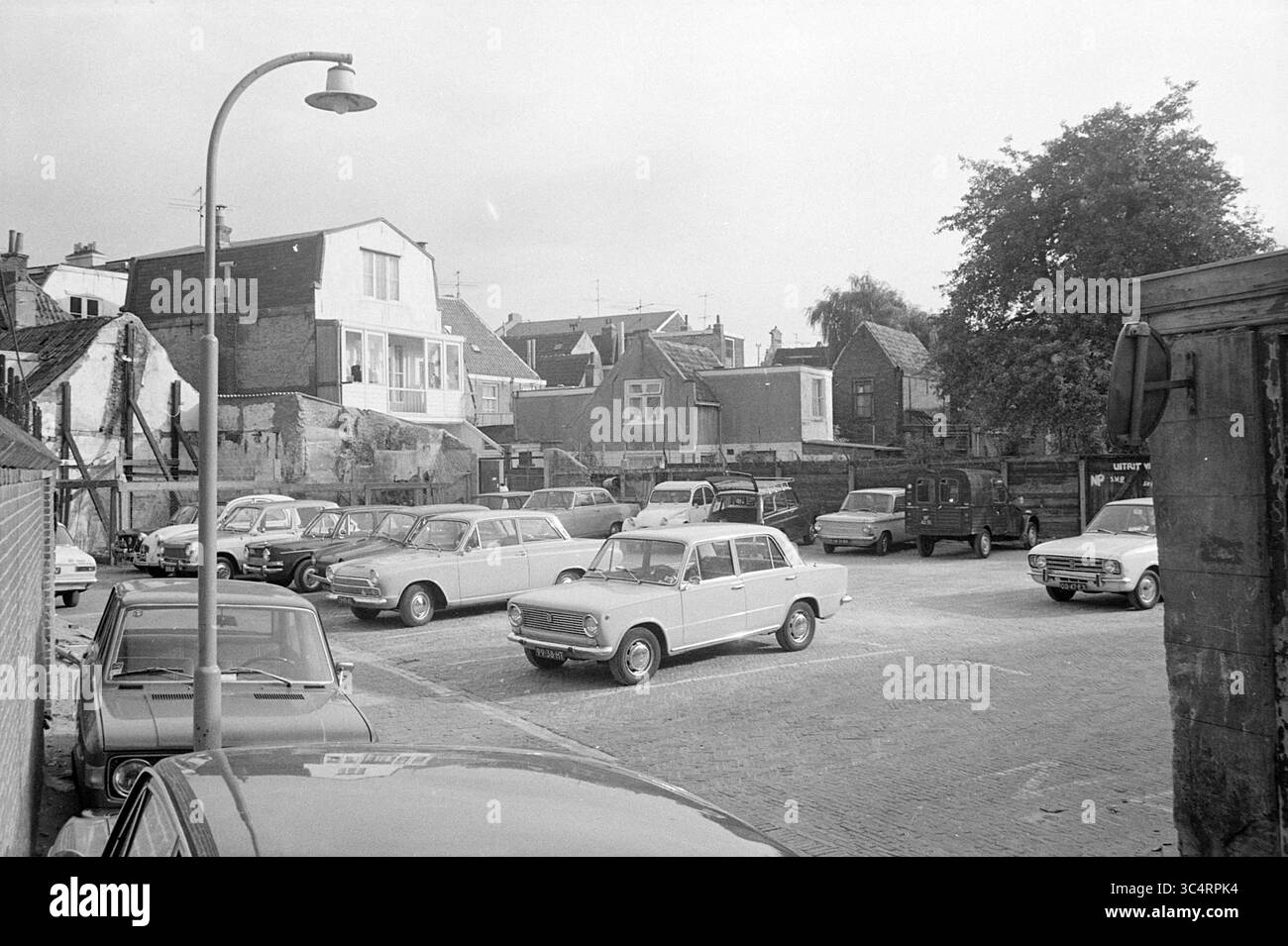 Parking with Cars Whizgle News, Dutch Desk, pays-Bas, 1950-2000 Un parking animé rempli de voitures anciennes, entouré d'un mélange de bâtiments résidentiels et commerciaux, présentant une tranche de la vie quotidienne d'une époque passée. Des arbres et un lampadaire ajoutent à la scène, faisant allusion à l'environnement urbain. Banque D'Images