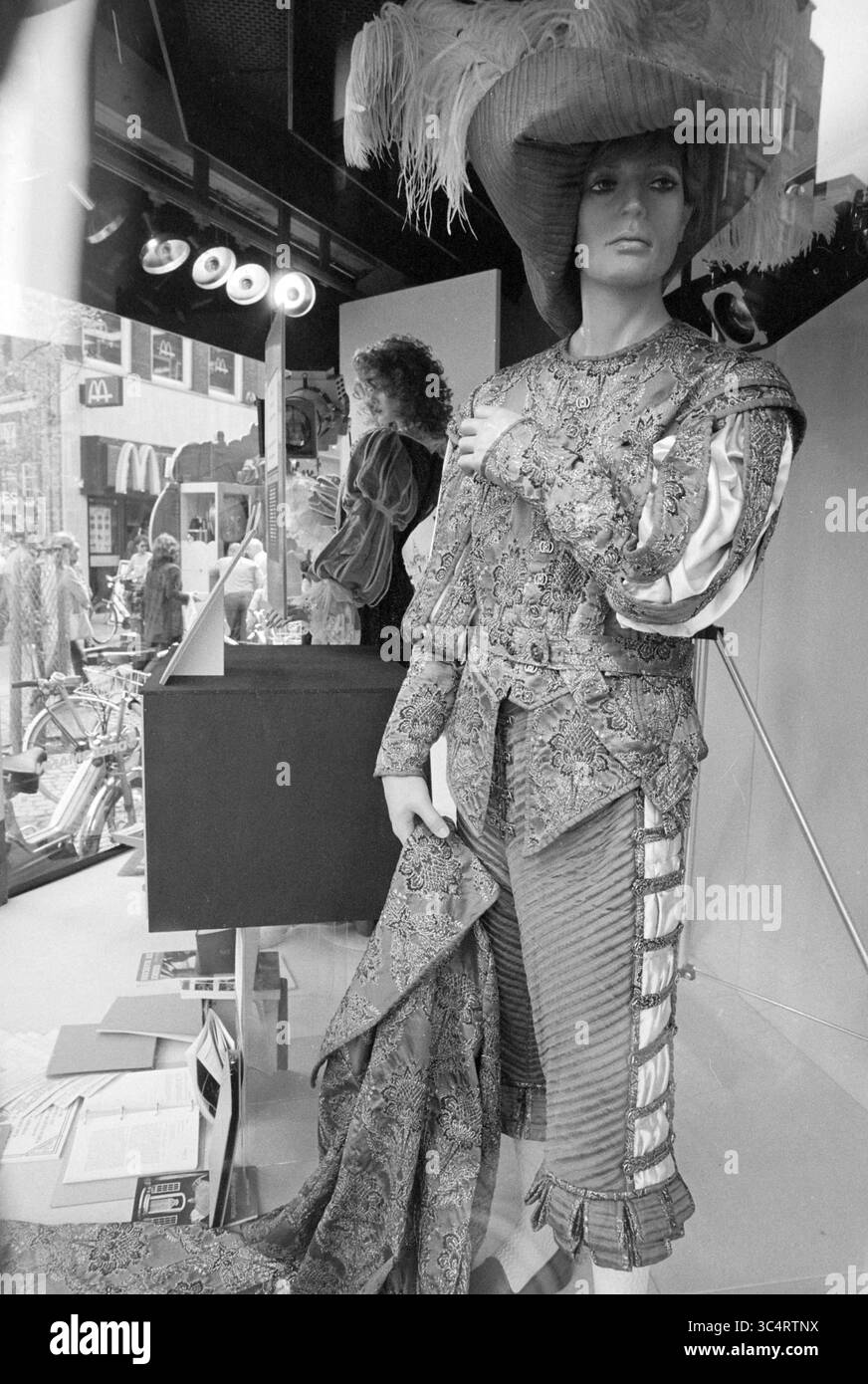 Mannequin en vitrine avec costume de scène, 09-05-1981 Whizgle News, Dutch Desk, pays-Bas, 1950-2000 Un mannequin élégant présente une tenue à motifs élaborés, avec un chapeau surdimensionné spectaculaire. En arrière-plan, une personne travaille derrière une exposition, ajoutant à l'atmosphère animée d'une rue avant-gardiste. Banque D'Images