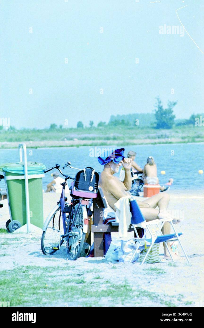 The Water test, Veerpolder, 21-08-1995 Whizgle News, Dutch Desk, pays-Bas, 1950-2000 Une scène détendue au bord de l'eau, mettant en scène un sunbather portant un chapeau à larges bords, assis confortablement sur une chaise à côté d'un vélo. À proximité, les gens profitent de la plage, avec quelques pataugeoires dans l'eau, tandis que des équipements de plage colorés ajoutent une touche vivante au cadre. Banque D'Images