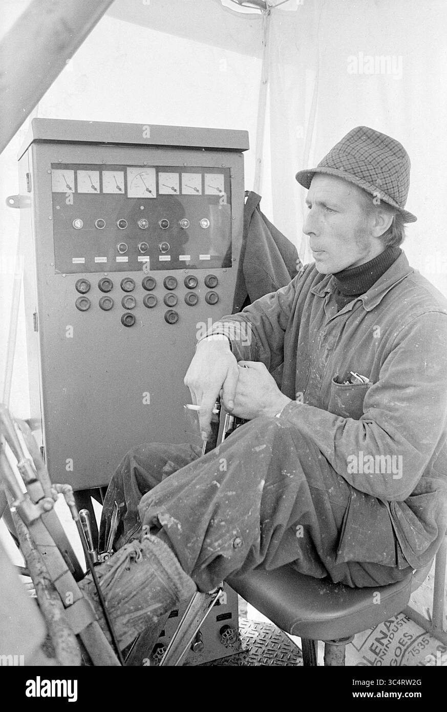 Pilote de pile Whizgle News, Dutch Desk, pays-Bas, 1950-2000 Un homme en tenue de travail s'assoit à un panneau de commande, intentionnellement concentré tout en faisant fonctionner des machines. Son chapeau et ses vêtements suggèrent un style robuste et pratique adapté à un environnement exigeant. Divers cadrans et boutons sont visibles sur le panneau, indiquant la complexité des tâches qu'il gère. Banque D'Images