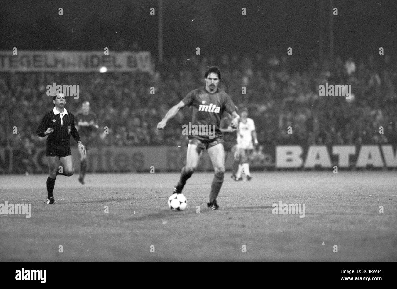 Football Haarlem - Ajax, 00-09-1983 Whizgle News, Dutch Desk, pays-Bas, 1950-2000 Un joueur de football dribble la balle en avant sur le terrain, tandis qu'un arbitre traîne derrière, observant le jeu. La scène capture l'intensité du match, avec une foule bondée visible en arrière-plan. Banque D'Images