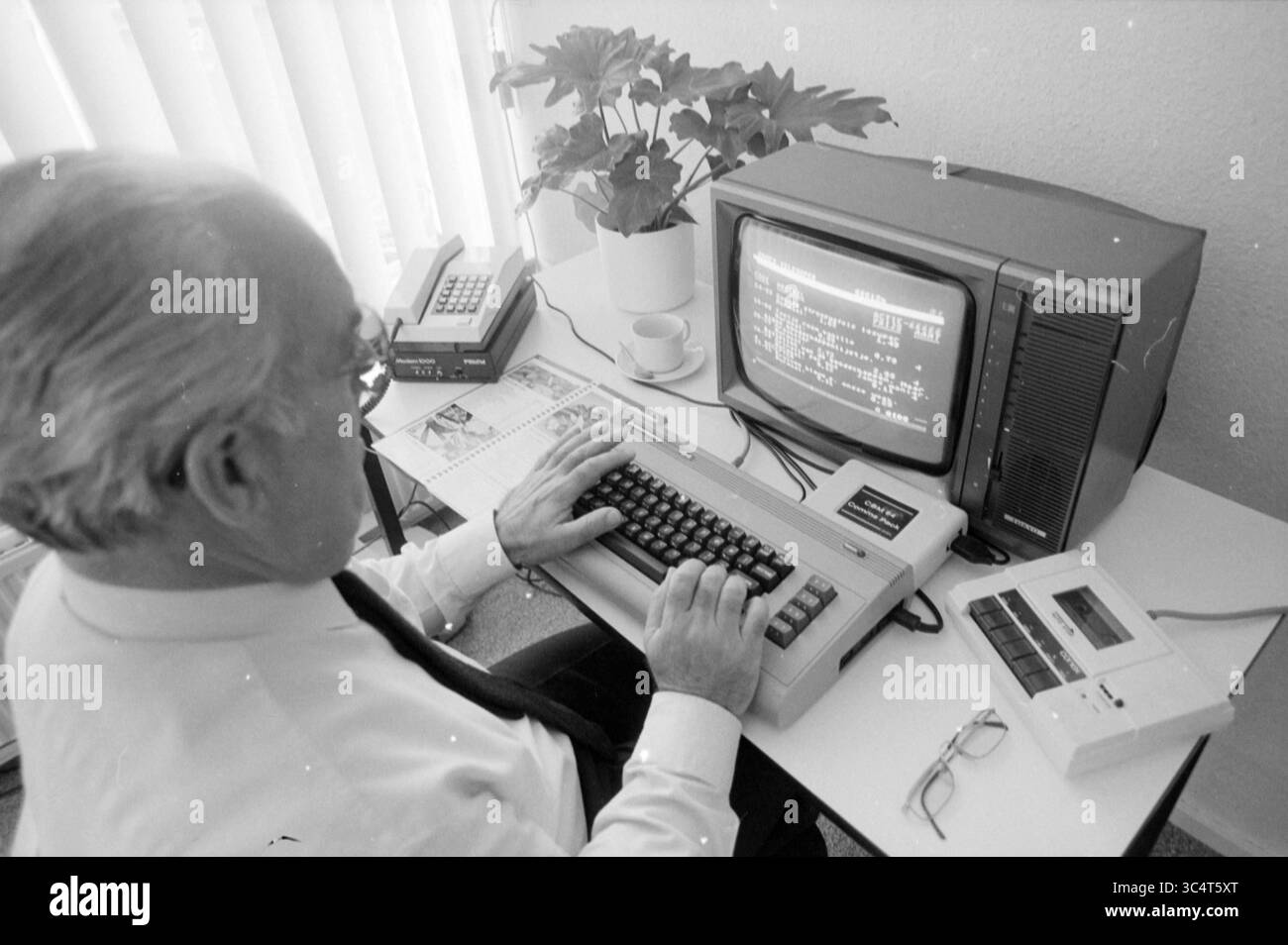 Réparation de centraux téléphoniques Mr. Beam, téléphone, téléphonie, 25-09-1987 Whizgle News, Dutch Desk, pays-Bas, 1950-2000 Un homme en costume est assis à un bureau, tapant sur un ordinateur vintage. L'écran affiche des lignes de code, tandis qu'un téléphone et un lecteur de cassettes sont situés à proximité, entourés de plantes en pot. Banque D'Images