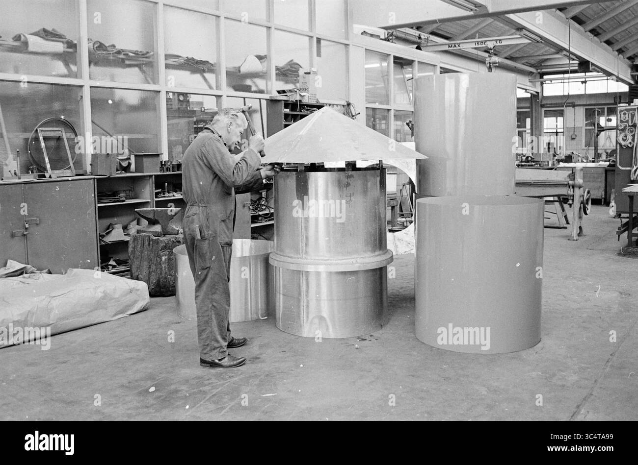 Mr. Willemse with Pipes, Persons, 22-09-1967 Whizgle News, Dutch Desk, pays-Bas, 1950-2000 Un artisan inspecte soigneusement une grande structure métallique cylindrique dans un atelier rempli d'outils et de matériaux. À proximité, diverses formes attendent un assemblage ultérieur, mettant en valeur un mélange de créativité et de design industriel. Banque D'Images