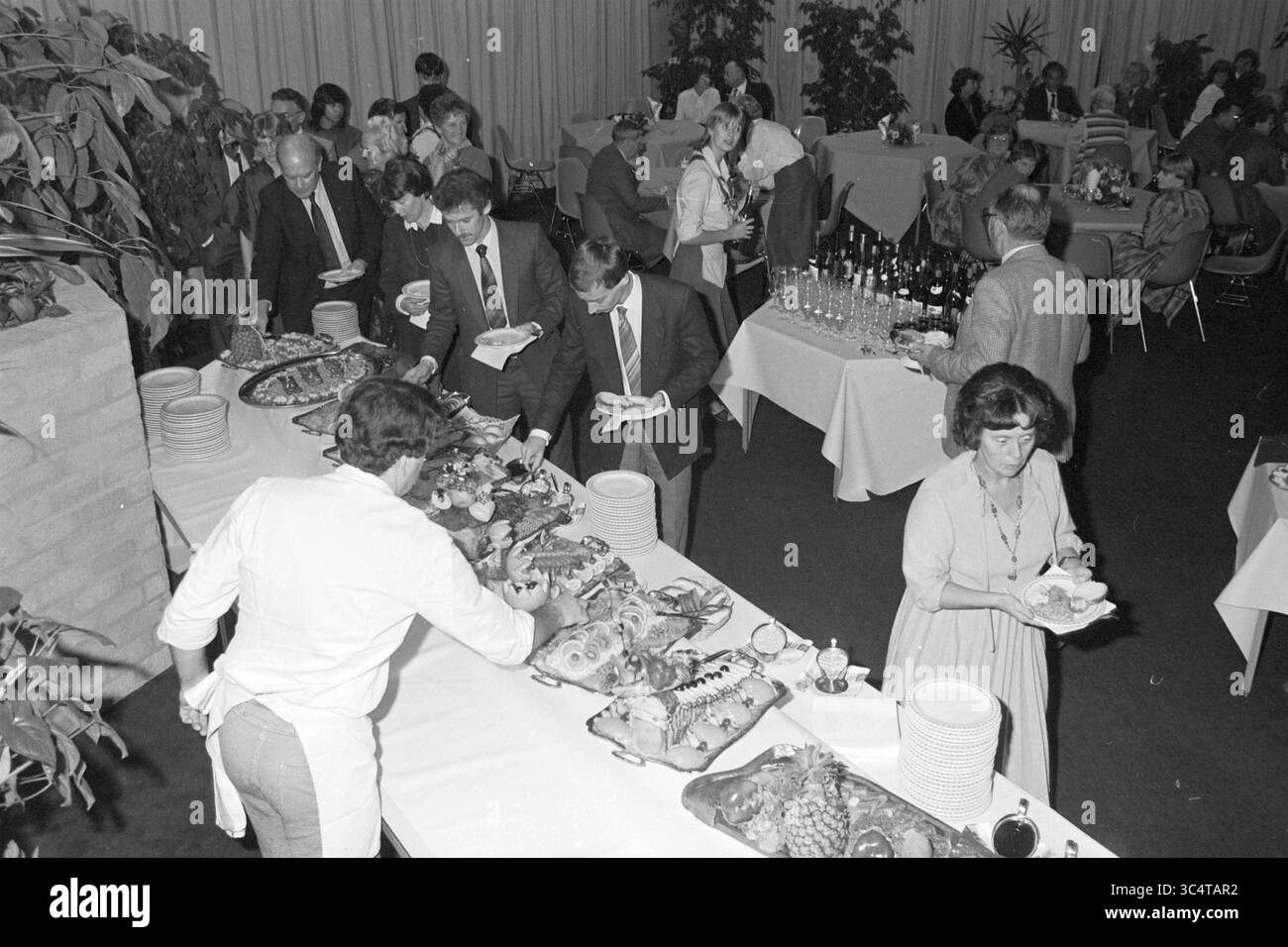 Grand espace de réception Whizgle News, Dutch Desk, pays-Bas, 1950-2000 Une scène de banquet animée avec les invités dégustant un buffet rempli d'une variété de plats. Les participants sont engagés dans la conversation, tandis que certains choisissent la nourriture d'une longue table ornée de plateaux de fruits de mer, fruits et autres délices. L'atmosphère est festive, avec des plantes décorant l'espace et des invités habillés en tenue formelle. Banque D'Images
