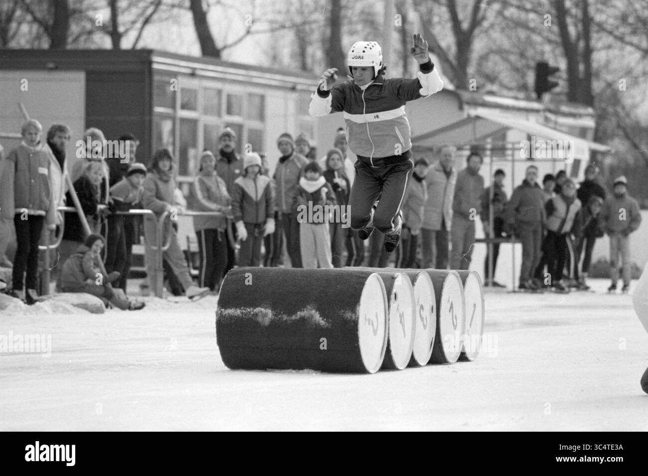 Patineurs sautant par-dessus quatre tonneaux sur une patinoire artificielle à Haarlem (« saut en tonneau »), Haarlem, pays-Bas, 09-02-1985 Whizgle News, Dutch Desk, pays-Bas, 1950-2000 un athlète saute habilement au-dessus d'un ensemble de grands barils sur une surface glacée, faisant preuve d'agilité et d'équilibre, tandis qu'une foule de spectateurs regarde en arrière-plan, regroupés contre le froid. Banque D'Images