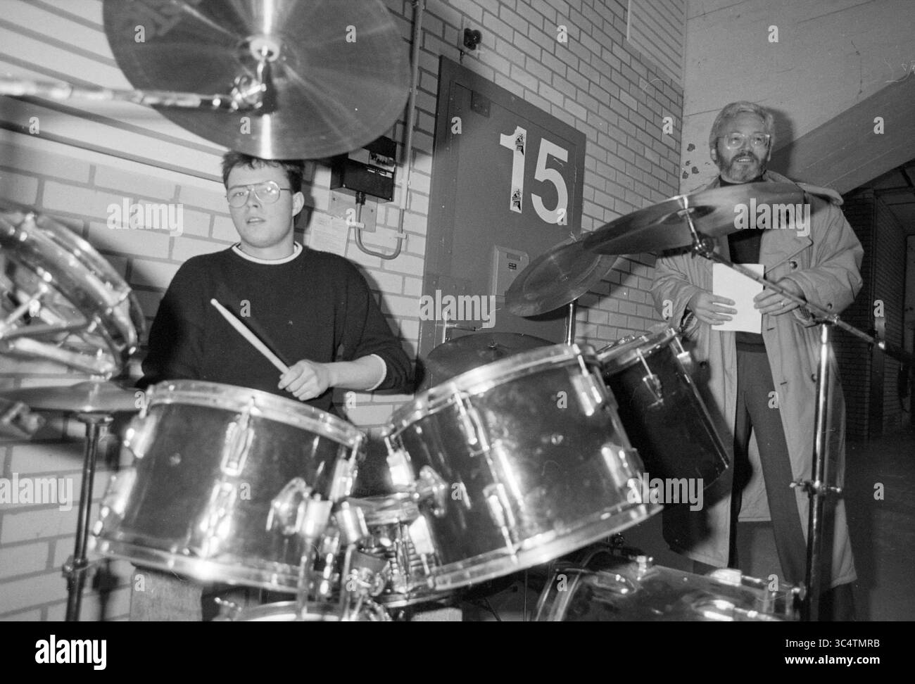 Ouverture POP bunker Kleijwegt., bunkers, ouverture, ouvertures, mise en service, 24-02-1989 Whizgle News, Dutch Desk, pays-Bas, 1950-2000 Un musicien s'entraîne sur une batterie tandis qu'une autre personne se tient à proximité, tenant un presse-papiers et observant la performance. L'environnement suggère un espace de répétition ou un studio, avec une porte numérotée visible en arrière-plan. Banque D'Images