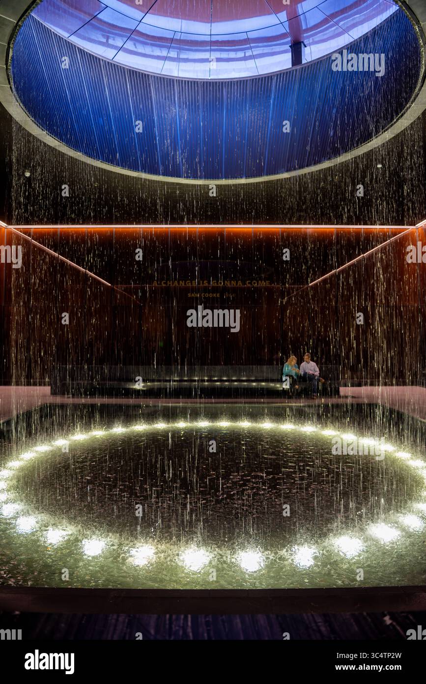 Musée national d'histoire et de culture afro-américaines fontaine de cour contemplative Washington DC | WASHINGTON D.C., États-Unis — Une vue verticale capture l'eau qui coule de l'oculus de la cour contemplative du Musée national d'histoire et de culture afro-américaines Smithsonian, encadrée par des murs de verre en cuivre-bronze. La cour occupe le niveau 0 du hall, fonctionnant comme un point de pause entre les galeries d'histoire du musée en dessous du niveau et les galeries culturelles au-dessus. Deux visiteurs sont assis sous une lyrique gravée de Sam Cooke, 'A change Is Gonna Come', l'une des nombreuses citations gravées dans t Banque D'Images