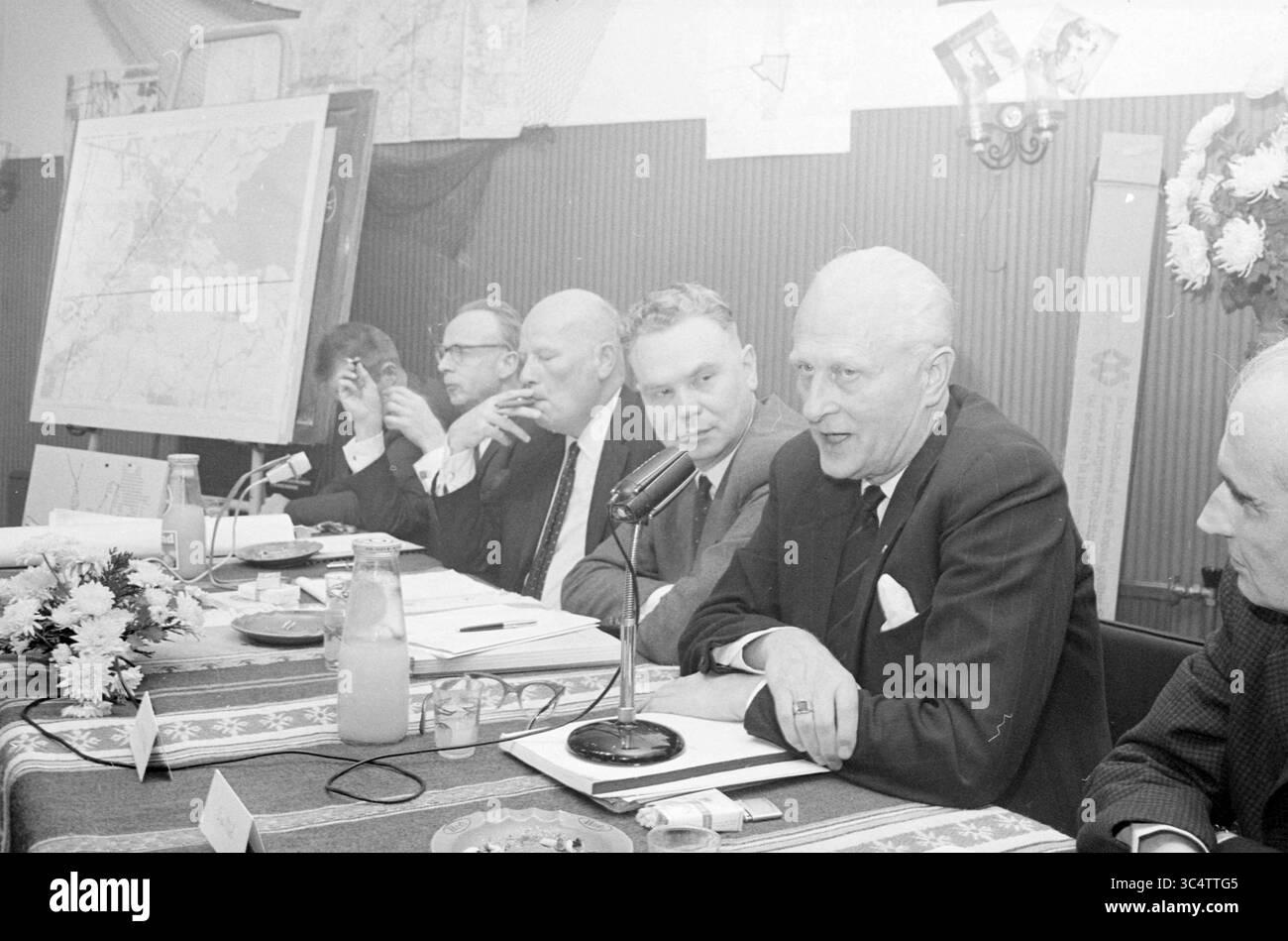 Teach-In Noise pollution Schiphol, Schiphol, 02-11-1967 Whizgle News, Dutch Desk, pays-Bas, 1950-2000 Un groupe de professionnels assis à une table s'engage dans une discussion formelle, avec des notes et des documents devant eux. Un homme parle dans un micro tandis que d'autres écoutent attentivement. Une grande carte est affichée en arrière-plan, suggérant un accent sur la planification ou la stratégie. Les éléments de décoration et les arrangements floraux ajoutent une touche de décorum au décor. Banque D'Images