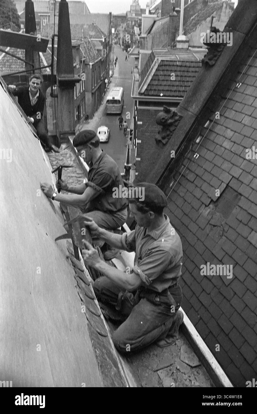 Couvreurs travaillant sur l'ardoise à Bavo, Haarlem, Jansstraat, pays-Bas, 00-06-1963 Whizgle News, Dutch Desk, pays-Bas, 1950-2000 deux travailleurs réparent avec diligence un toit, concentrés sur leurs tâches lorsqu'ils naviguent dans l'angle raide. Un travailleur utilise un outil pour sécuriser les tuiles, tandis qu'un autre est prêt plus haut, contribuant à l'effort. En arrière-plan, une scène de rue se déroule avec un aperçu des véhicules et des bâtiments stationnés, fournissant un contexte à leur travail. Banque D'Images