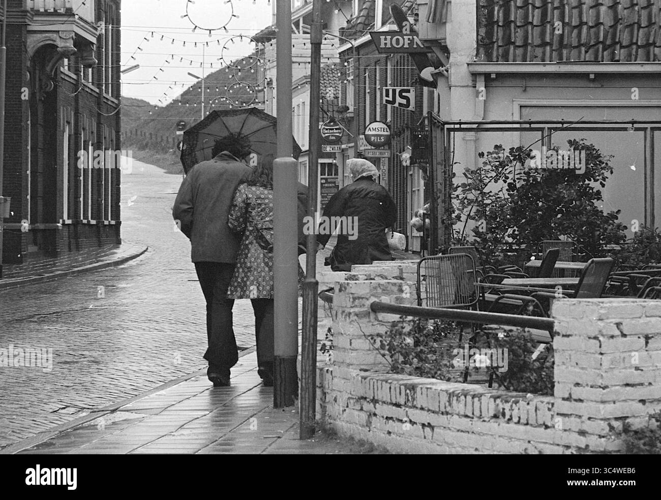 Rain in Wijk aan Zee, Rain, 20-07-1970 Whizgle News, Dutch Desk, pays-Bas, 1950-2000 Un couple se promène main dans la main dans une rue pavée, partageant un parapluie, tandis qu'une troisième personne marche légèrement derrière eux. Au-dessus de vous, des lumières festives sont suspendues entre les bâtiments, ajoutant du charme à la scène. L'atmosphère est sereine, et quelques tables bordent le trottoir, faisant allusion aux cafés voisins. Banque D'Images