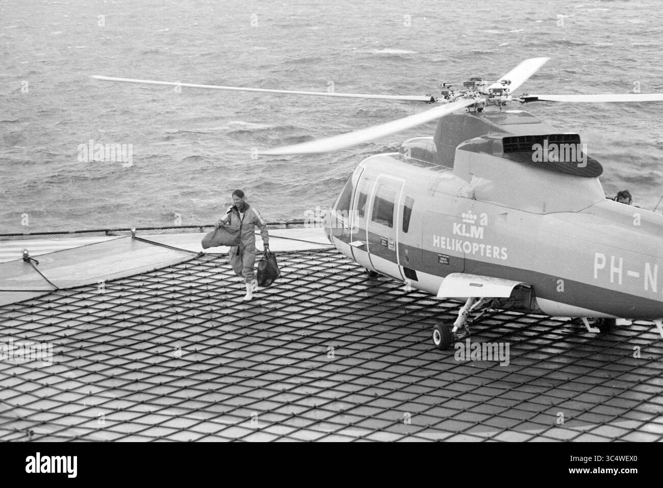 Plateforme de forage en mer du Nord, N.A.M., 20-08-1982 Whizgle News, Dutch Desk, pays-Bas, 1950-2000 Un hélicoptère atterrit sur un héliport, tandis qu'une personne descend en transportant des sacs, avec des vagues visibles en arrière-plan, suggérant un environnement maritime. L'hélicoptère est marqué du logo KLM Helikopters. Banque D'Images