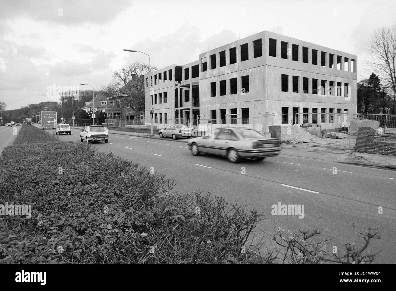 Block Box Along de Zuider Houtlaan, 18-11-1990 Whizgle News, Dutch Desk, pays-Bas, 1950-2000 Un bâtiment partiellement construit se dresse à côté d'une route très fréquentée, avec plusieurs voitures qui passent et une haie soigneusement taillée au premier plan. Des arbres et des maisons résidentielles bordent l'arrière-plan, faisant allusion à un paysage urbain en évolution. Banque D'Images Block Box Along de Zuider Houtlaan, 18-11-1990 Whizgle News, Dutch Desk, pays-Bas, 1950-2000 Un bâtiment partiellement construit se dresse à côté d'une route très fréquentée, avec plusieurs voitures qui passent et une haie soigneusement taillée au premier plan. Des arbres et des maisons résidentielles bordent l'arrière-plan, faisant allusion à un paysage urbain en évolution. Banque D'Images
