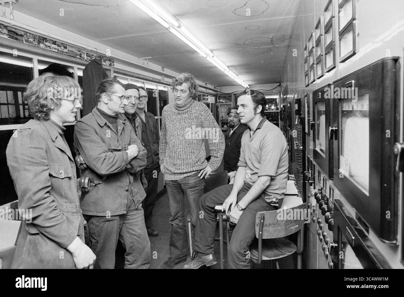 International Usine de machines à Hoogovens Whizgle News, Dutch Desk, pays-Bas, 1950-2000 Un groupe de six personnes s'engage dans la conversation, rassemblées autour de panneaux de commande remplis d'écrans et de cadrans. Le cadre semble être un espace de travail ou une installation technique, avec des expressions ciblées et une atmosphère décontractée au sein du groupe. Banque D'Images