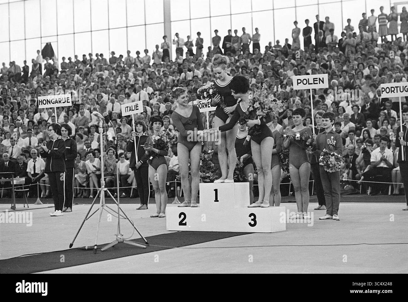 Championnats d'Europe de gymnastique artistique Amsterdam le 25/26 mars 1967, Amsterdam, Nederland, 25-03-1967 Whizgle News, Dutch Desk, pays-Bas, 1950-2000 trois athlètes se tiennent fièrement sur un podium, célébrant leurs réalisations lors d'un événement compétitif. La gagnante de la première place rayonne de joie, tandis que les concurrents de la deuxième et de la troisième place l'embrassent dans la camaraderie. Un large public remplit l'arrière-plan, avec divers panneaux indiquant leurs nationalités. Banque D'Images
