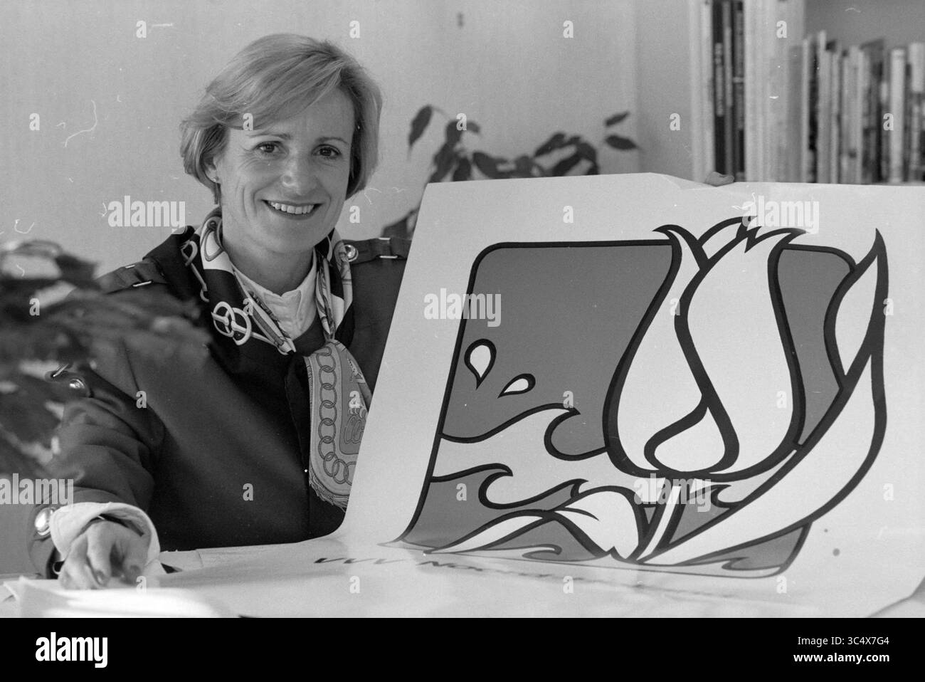 Affiche 'VVV Noord-Holland' avec M. van Bodegraven et le personnel provincial House, 16-01-1991 Whizgle News, Dutch Desk, pays-Bas, 1950-2000 Une femme est assise à une table, souriante en présentant un grand design avec une fleur stylisée et des formes fluides, entourée de livres et de plantes. Banque D'Images