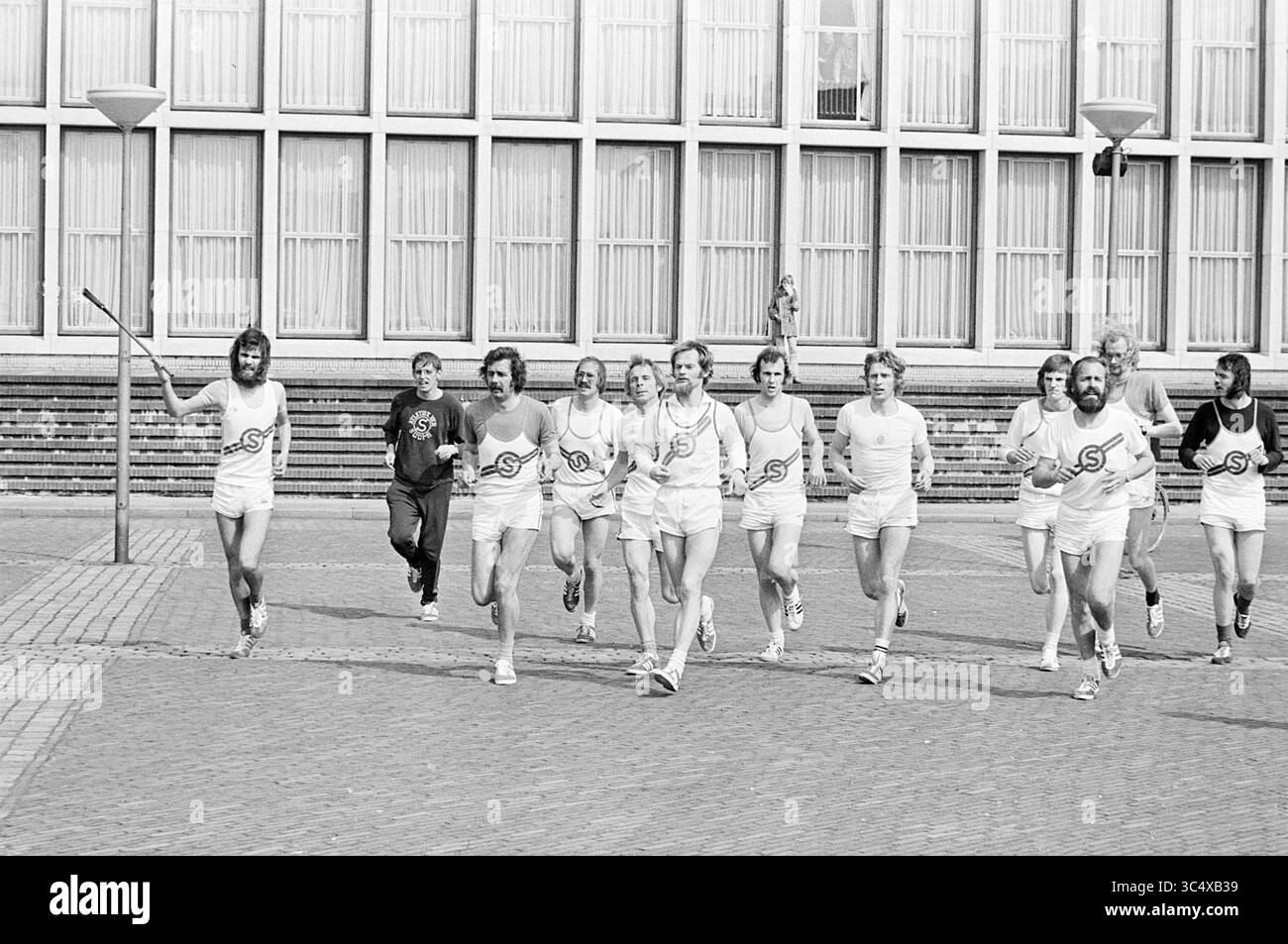5 mai célébration Velsen Torch run pseudo Canadiens, libération, jour de la libération, célébrations de la libération, 05-05-1975 Whizgle News, Dutch Desk, pays-Bas, 1950-2000 Un groupe de coureurs en tenue athlétique jogging ensemble le long d'une passerelle pavée, faisant preuve de camaraderie et de travail d'équipe. Au premier plan, un participant agite énergiquement un bras, tandis qu'un autre coureur traîne derrière, ajoutant à la scène animée. La toile de fond présente une série de grandes fenêtres, faisant allusion à un bâtiment à proximité. Banque D'Images