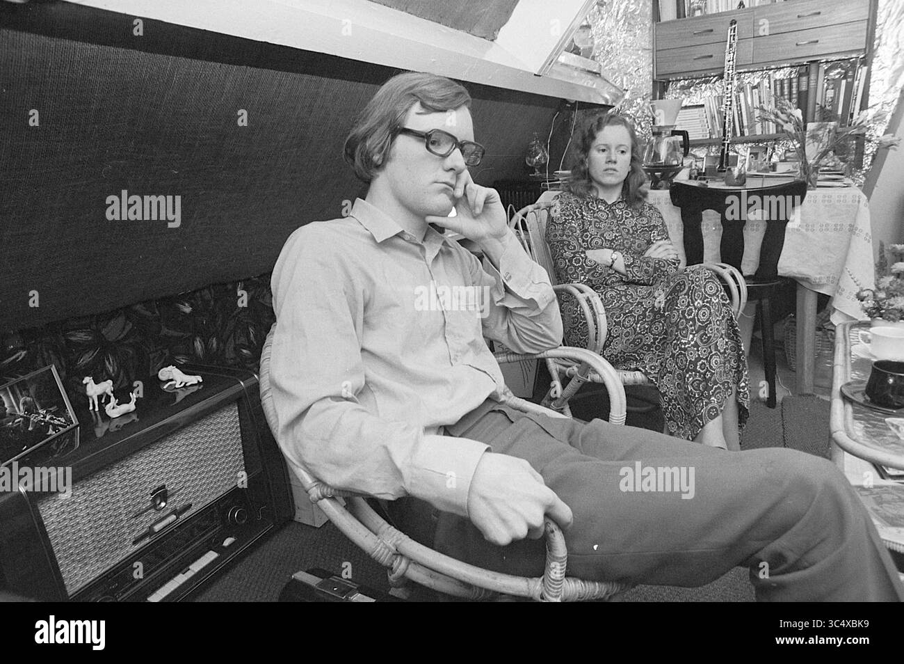 Un couple dans leur maison, 00-00-1972 Whizgle News, Dutch Desk, pays-Bas, 1950-2000 Un homme est assis avec soin dans une chaise en rotin, une main sur son menton, tandis qu'une femme dans une robe à motifs est assise à proximité, semblant contemplative. Le cadre comprend un décor vintage, des livres sur une étagère et une radio à l'ancienne, créant une atmosphère nostalgique. Banque D'Images