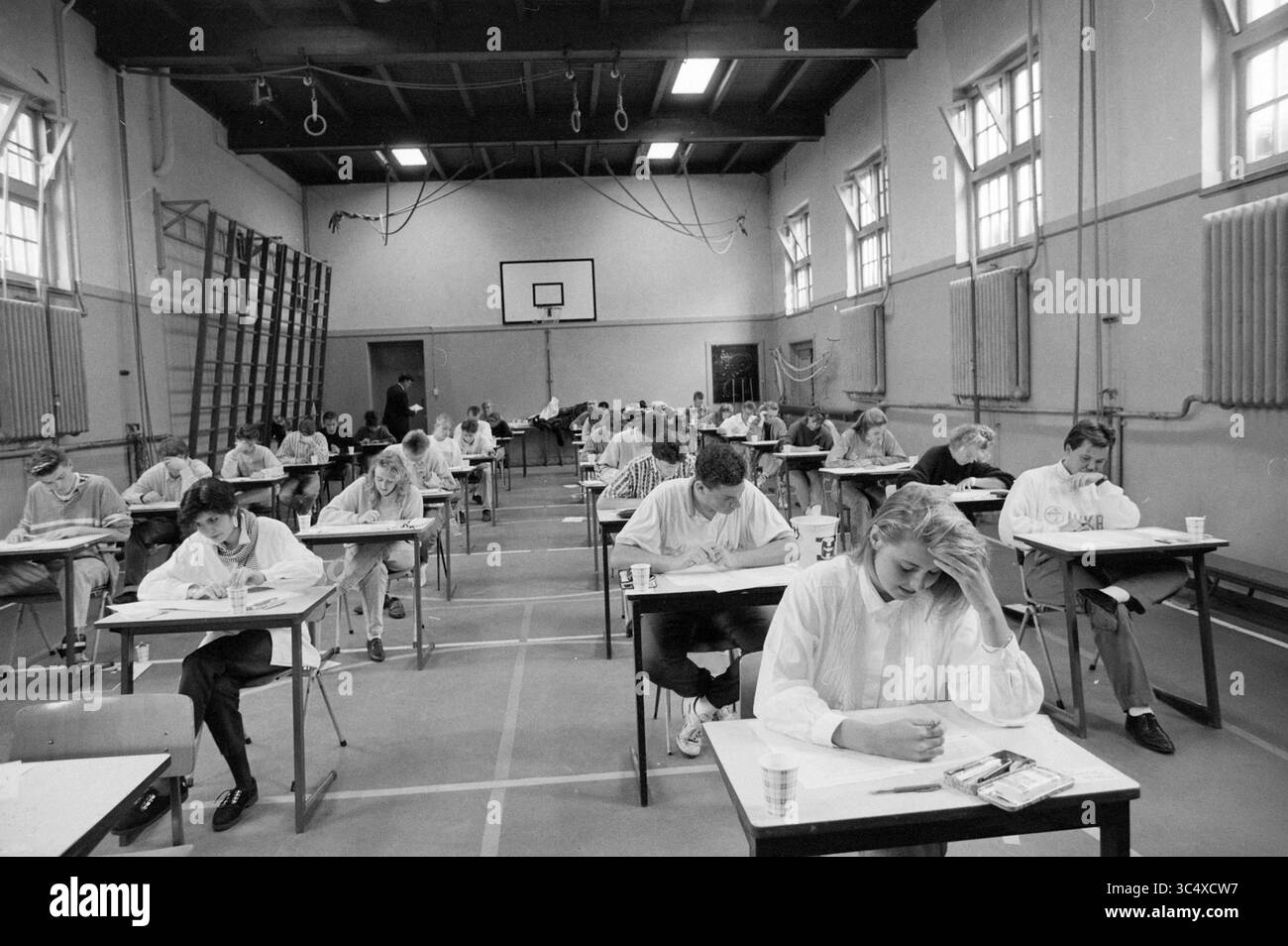 Examen final Trinity Lyceum, 14-05-1990 Whizgle News, Dutch Desk, pays-Bas, 1950-2000 étudiants sont assis à des bureaux dans un gymnase, axés sur leurs examens. L’atmosphère est tendue lorsque les individus se concentrent, certains montrant des signes de stress tandis que d’autres écrivent diligemment, entourés de grandes fenêtres qui laissent entrer la lumière naturelle. Banque D'Images
