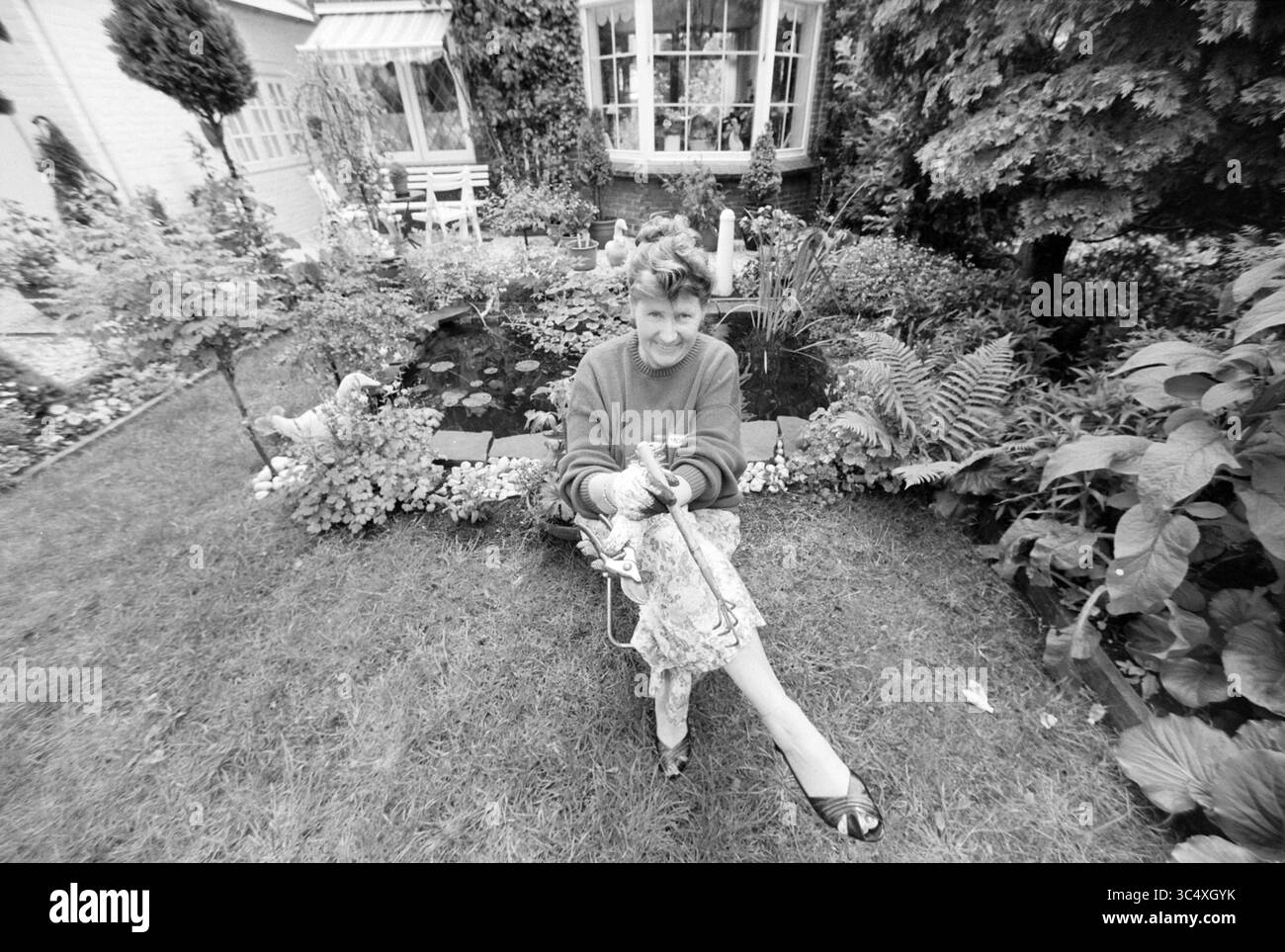 MRS Jansen, Garden Pride, 25-06-1991 Whizgle News, Dutch Desk, pays-Bas, 1950-2000 Une personne est assise de manière ludique dans un jardin luxuriant, entouré d'une verdure vibrante et d'un petit étang. Ils portent un pull lumineux et tiennent un morceau de tissu coloré, souriant joyeusement comme ils interagissent avec leur environnement. Banque D'Images
