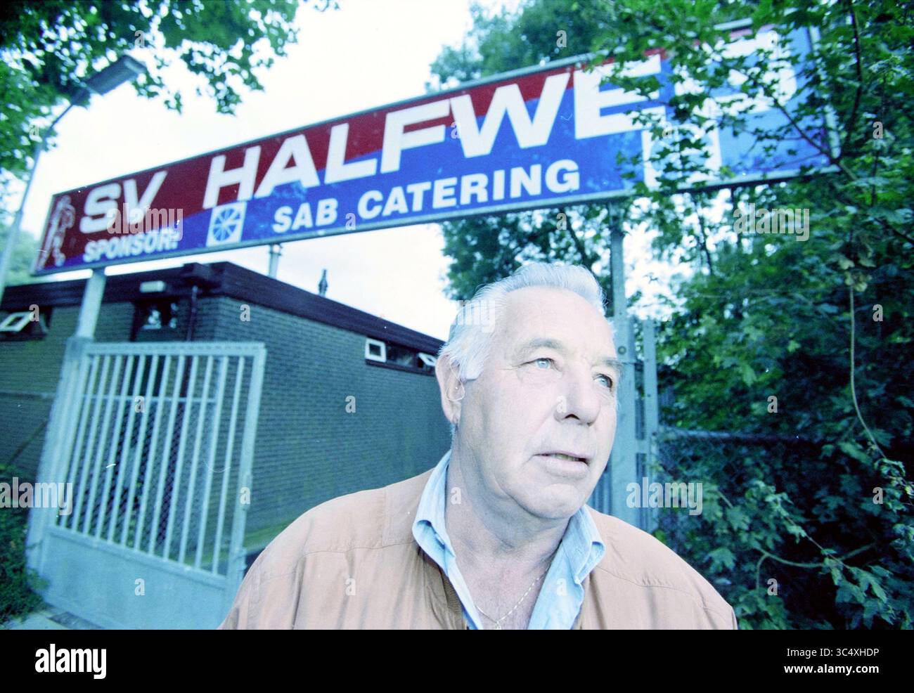 Dirk Vaandering Zwanenburg, Zwanenburg, 09-09-2002 Whizgle News, Dutch Desk, pays-Bas, 1950-2000 Un homme se tient devant un grand panneau pour SV Halfwet, un club sportif, avec les détails du parrainage visibles. Les environs comprennent de la verdure et une clôture, soulignant une atmosphère accueillante mais nostalgique. Banque D'Images