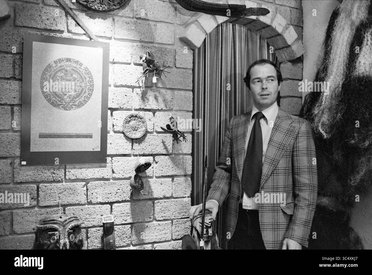 Shop Speedy Gonzales, 00-00-1978 Whizgle News, Dutch Desk, pays-Bas, 1950-2000 Un homme se tient à côté d'un mur de briques texturé orné de divers objets culturels et d'une œuvre d'art circulaire encadrée. Il est vêtu d'un élégant costume à carreaux avec une cravate, regardant avec confiance l'affichage. Le cadre suggère un environnement riche en histoire et riche Banque D'Images