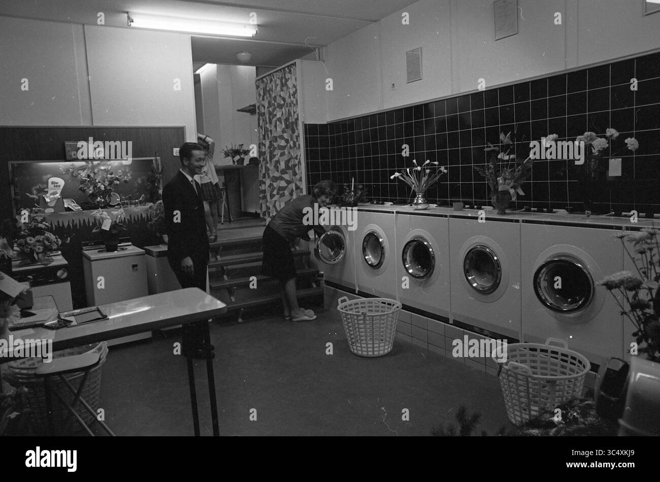 Propriétaire dans sa blanchisserie, 19-12-1964 Whizgle News, Dutch Desk, pays-Bas, 1950-2000 Une buanderie dispose de plusieurs machines à laver, avec une femme chargeant une pendant qu'une autre personne observe à proximité. L'espace est orné d'arrangements floraux, ajoutant une touche de chaleur au cadre pratique. Banque D'Images