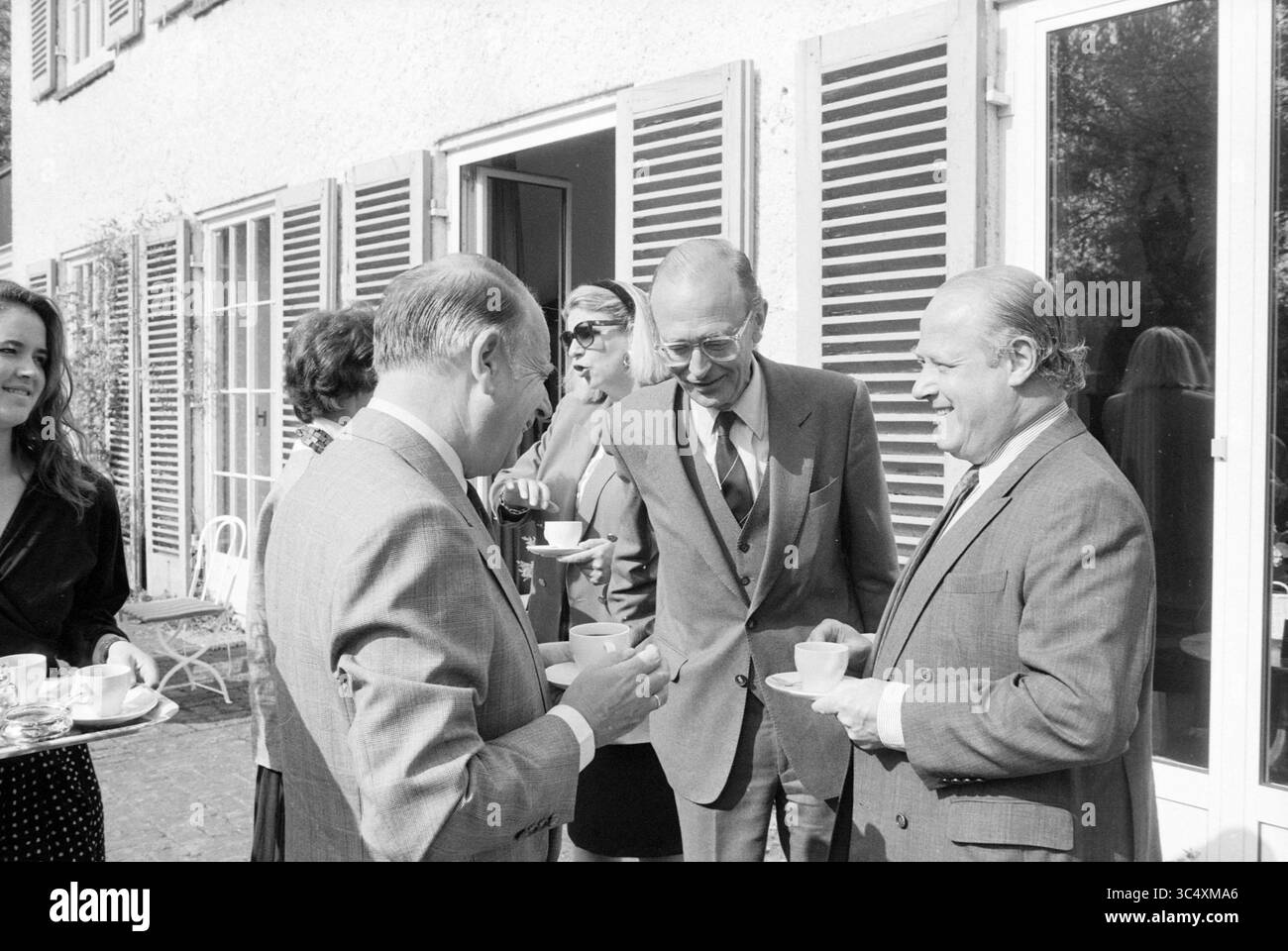 Une réception pour l'honneur royal de M. Fronestius, Bloemendaal, directeur d'ABN Whizgle News, Dutch Desk, pays-Bas, 1950-2000 Un groupe d'individus élégamment habillés s'engage dans la conversation, chacun tenant une tasse, alors qu'ils profitent d'un rassemblement social en plein air. L’ambiance est détendue, avec en toile de fond des portes partiellement ouvertes et des volets, suggérant un cadre agréable pour l’événement. Banque D'Images