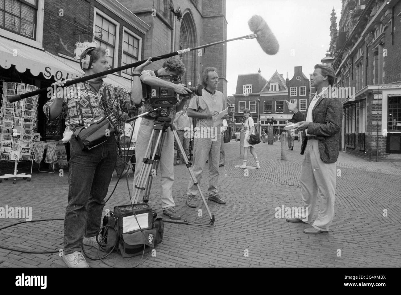 Enregistrements TV avec Willem Nijholt, 15-08-1991 Whizgle News, Dutch Desk, pays-Bas, 1950-2000 Une équipe de tournage capture un reportage en direct sur une place urbaine animée, où un présentateur de nouvelles est prêt à livrer un segment, flanqué de caméramans et de techniciens audio. Autour d'eux, l'architecture pittoresque des magasins et des bâtiments ajoute une charmante toile de fond à la scène. Banque D'Images