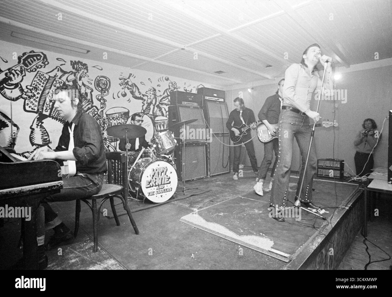 Long Tall Ernie et les Shakers performance Whizgle News, Dutch Desk, pays-Bas, 1950-2000 Un groupe dynamique se produit sur scène, avec un chanteur chantant passionnément dans un microphone tandis qu'un pianiste joue à proximité. La scène est animée, avec un batteur et un guitariste, et se déroule sur fond de mur coloré L'énergie de la performance captive le public, créant une atmosphère engageante. Banque D'Images