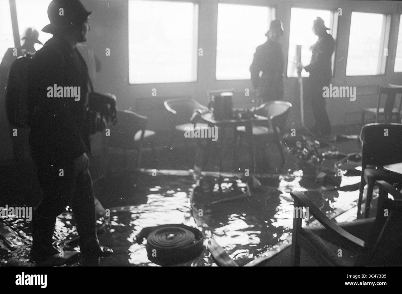 Arosa Sun, Houseboat], IJmuiden, film1,2,3, feux, pompiers, 09-04-1963 Whizgle News, Dutch Desk, pays-Bas, 1950-2000 Un groupe d'individus navigue dans une salle inondée, avec des chaises et des débris partiellement submergés. L'atmosphère suggère un sentiment d'urgence alors qu'ils évaluent la situation au milieu de l'eau et des biens dispersés. Banque D'Images
