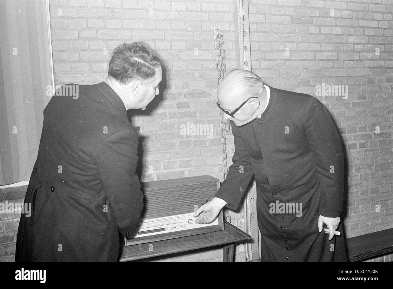 Un ecclésiastique et un autre homme à une radio Whizgle News, Dutch Desk, pays-Bas, 1950-2000 deux hommes en tenue formelle examinent et discutent d'une radio vintage ou d'un équipement audio, l'un ajustant un cadran tandis que l'autre observe attentivement. Le cadre présente des murs de briques apparentes, suggérant un environnement historique ou institutionnel. Banque D'Images