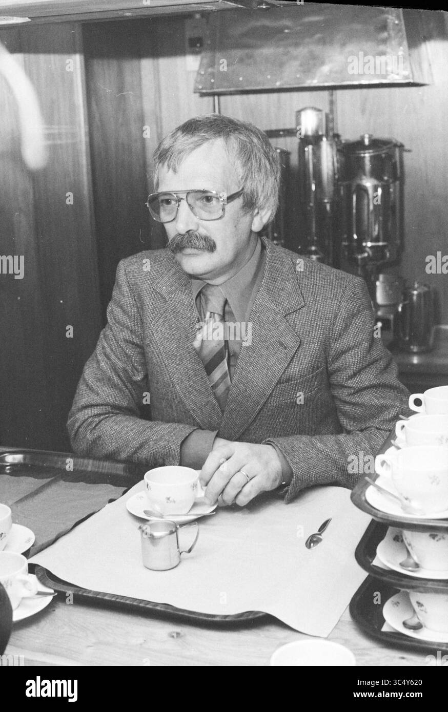 Diverses personnes en conversation, 07-05-1979 Whizgle News, Dutch Desk, pays-Bas, 1950-2000 Un homme avec une moustache et des lunettes est assis à une table, vêtu d'un costume avec une cravate à motifs. Une tasse et une soucoupe sont positionnées devant lui, ainsi qu'une petite crémeuse. Le décor présente une toile de fond chaude en bois et un aperçu d'un distributeur de boissons en arrière-plan. Banque D'Images