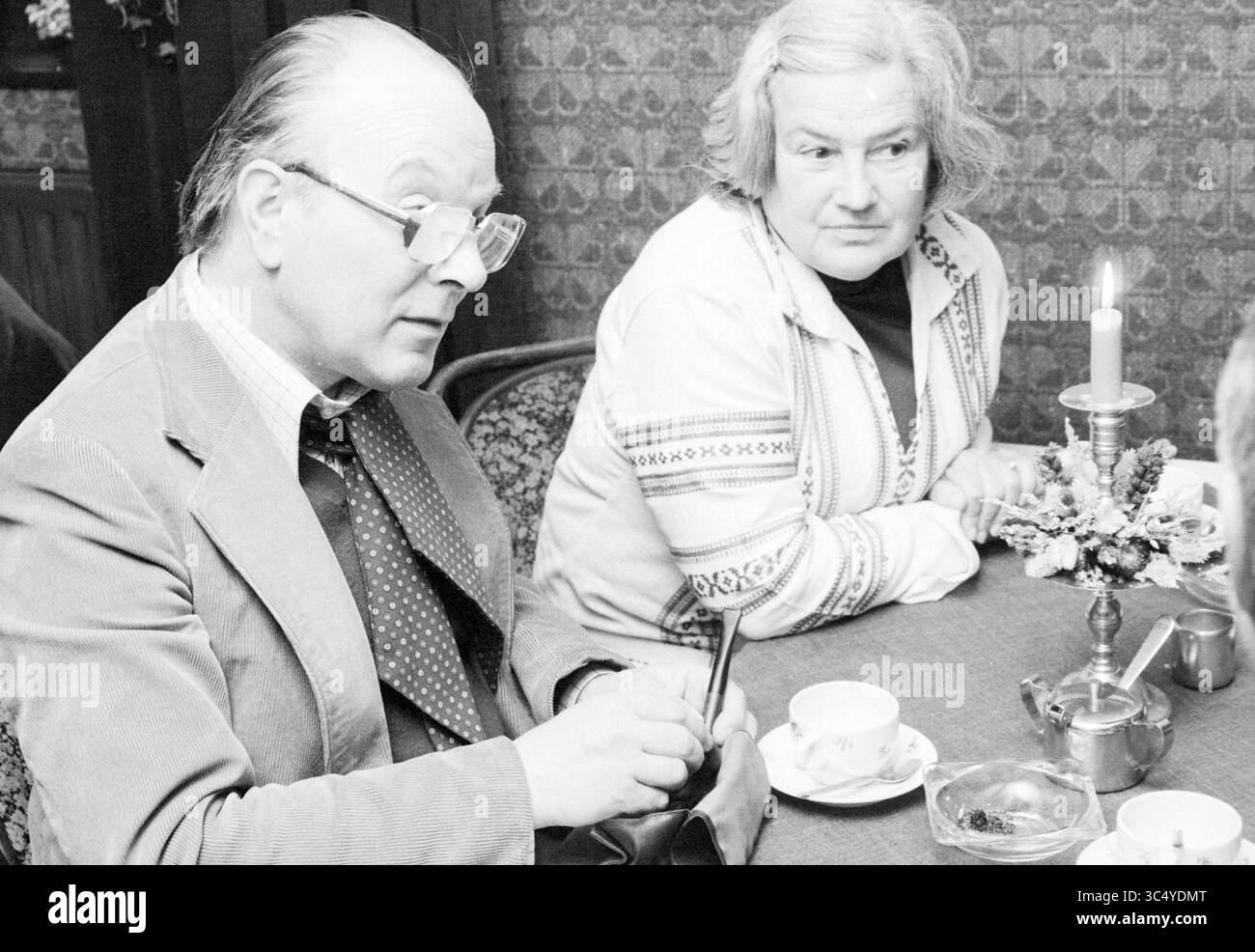 Diverses personnes en conversation, 07-05-1979 Whizgle News, Dutch Desk, pays-Bas, 1950-2000 Un homme en costume et lunettes est assis à une table, concentré sur l'écriture, tandis qu'une femme avec une coiffure de couleur claire regarde soigneusement à côté de lui. La table est sertie de tasses, d'une bougie décorative et de petits arrangements floraux, créant une atmosphère chaleureuse pour la conversation. Banque D'Images