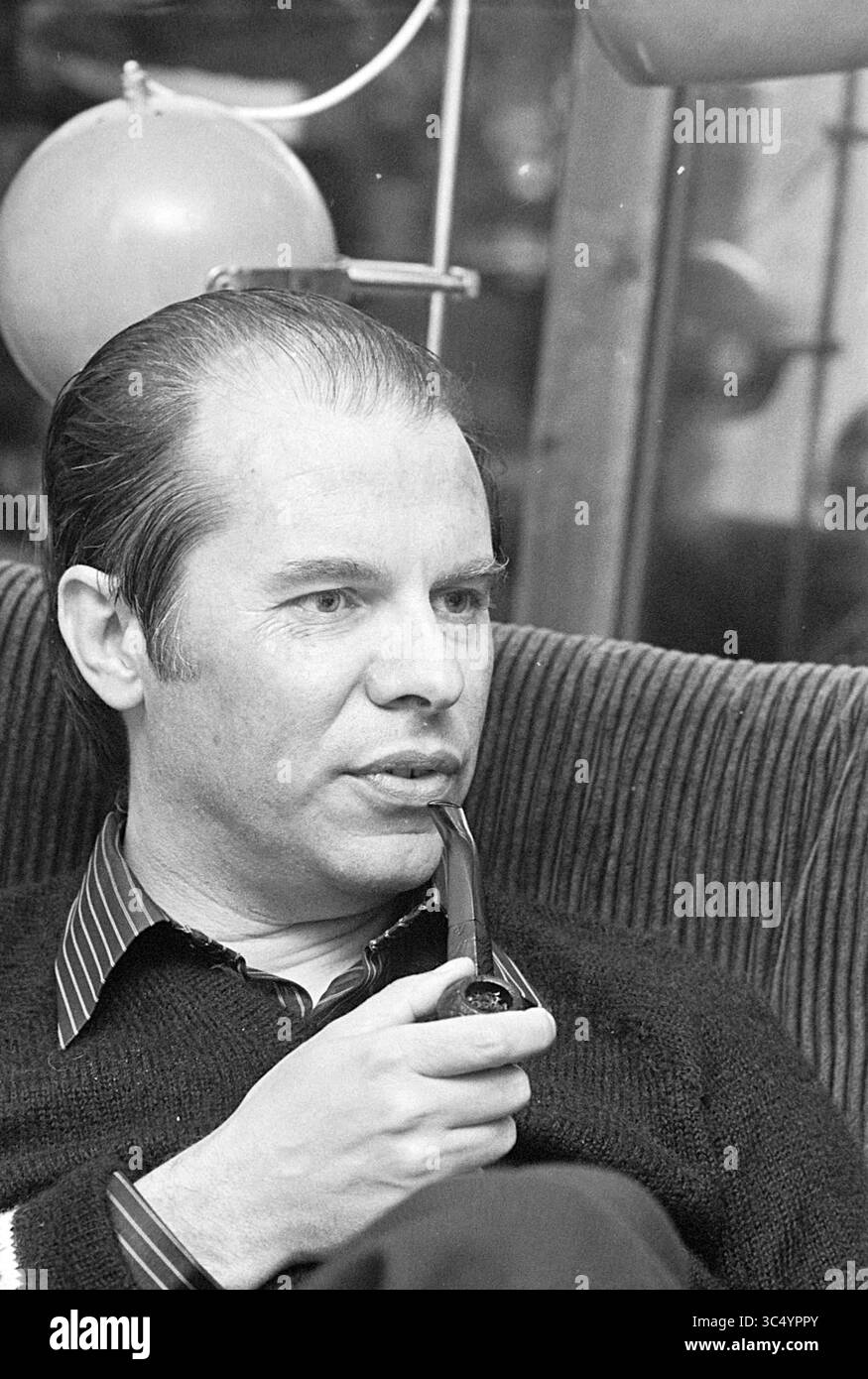 Portrait Mr. Van Geet in Santpoort + Mrs. T. Bolland te Driehuis., Portraits, Portraits, 12-05-1975 Whizgle News, Dutch Desk, pays-Bas, 1950-2000 Un homme est assis avec soin dans un cadre confortable, tenant une pipe et vêtu d'un pull tricoté sur une chemise à col. Il y a une expression contemplative sur son visage, suggérant qu'il est profondément dans la pensée ou engagé dans la conversation. Les ballons et les tissus d'ameublement en arrière-plan laissent entrevoir une atmosphère détendue. Banque D'Images