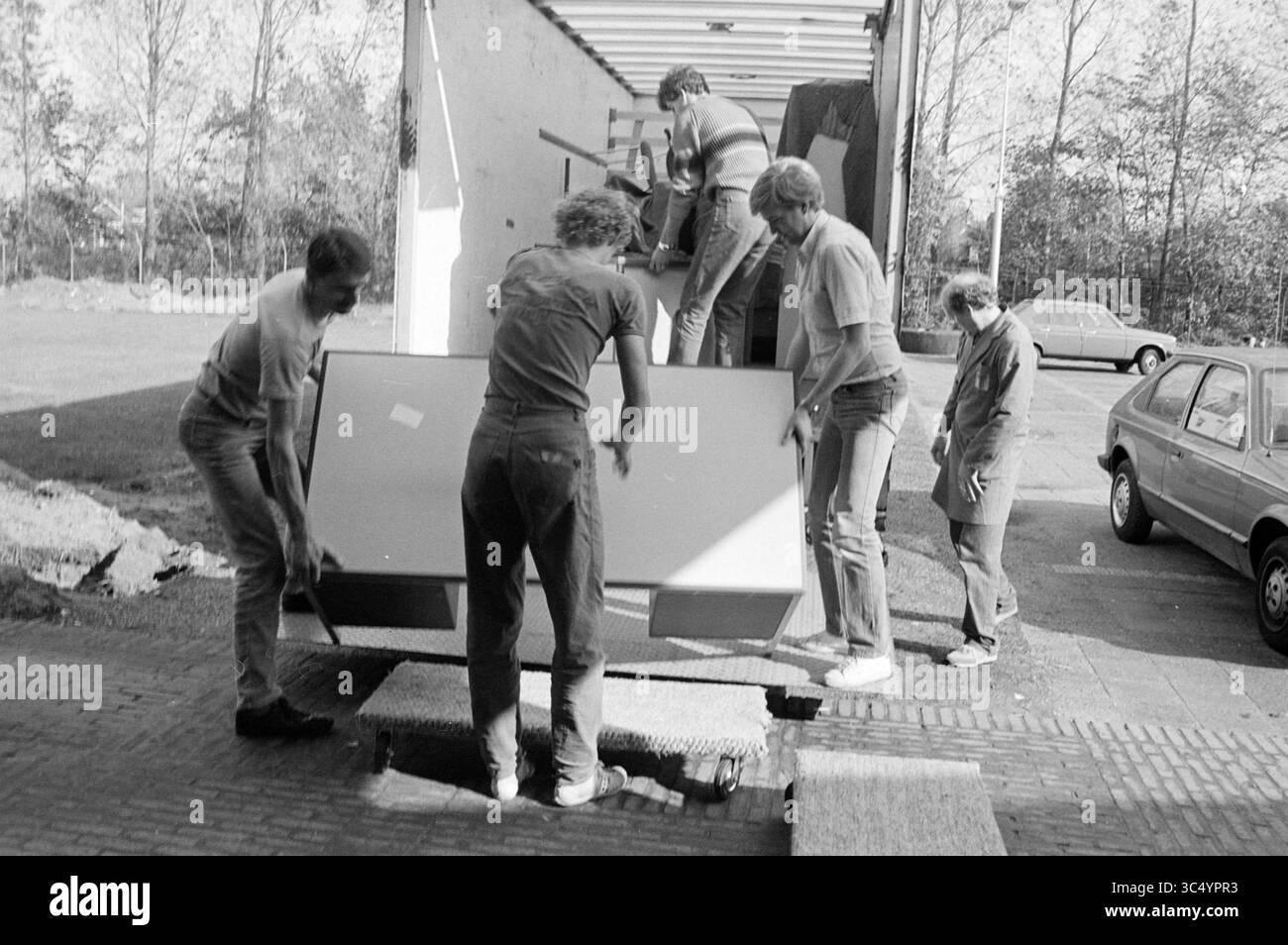 Déménagement à Velsen, N.A.M., Velsen, 16-10-1983 Whizgle News, Dutch Desk, pays-Bas, 1950-2000 Un groupe de cinq hommes décharge soigneusement les gros objets d'un camion de livraison, faisant preuve de travail d'équipe alors qu'ils gèrent divers meubles et équipements dans une aire de stationnement. A proximité, une voiture garée est visible, et des arbres peuvent être vus en arrière-plan, suggérant une journée calme. Banque D'Images