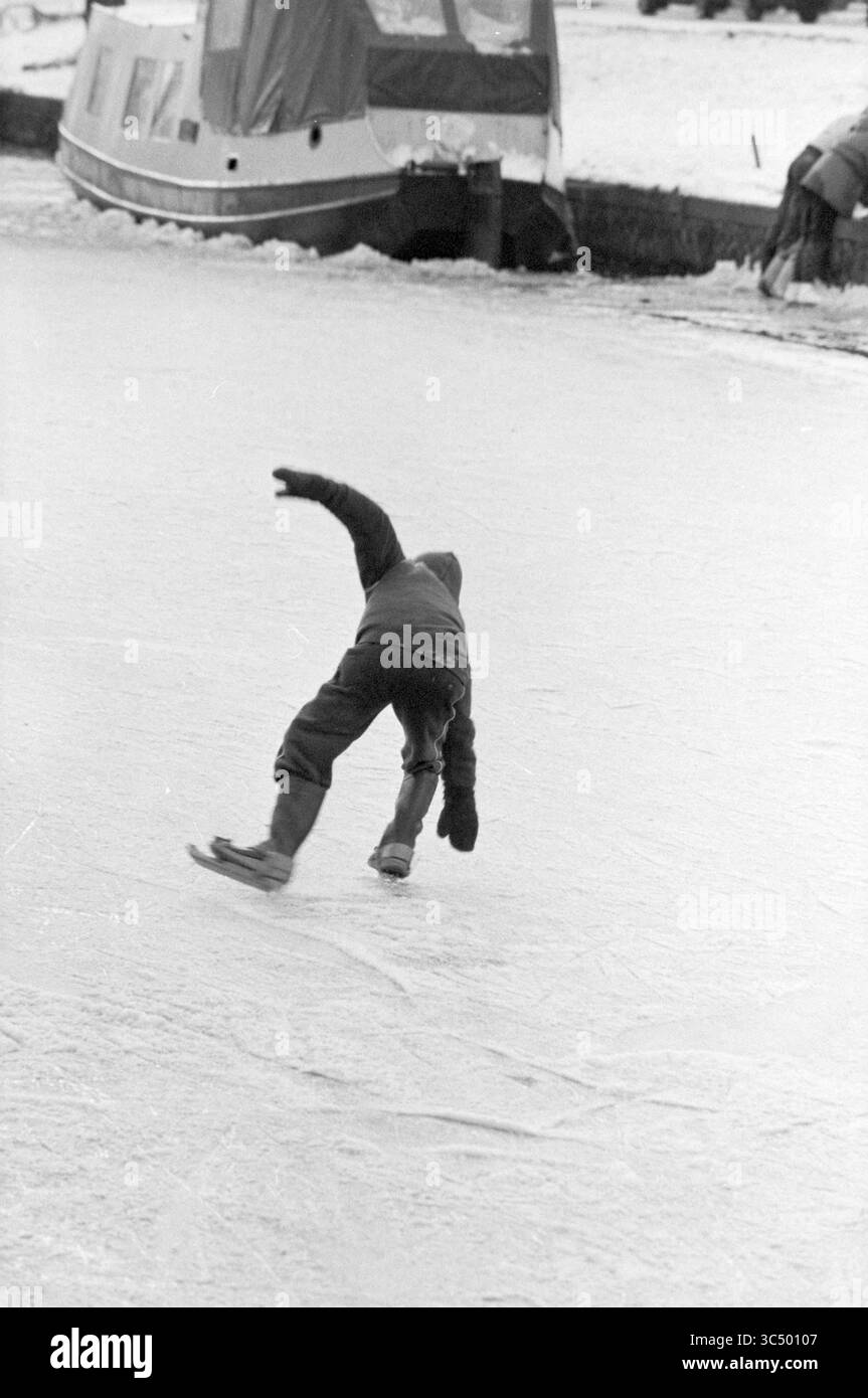Patinage sur glace à Kinderhuissingel, patinage sur glace, Haarlem, Kinderhuissingel, pays-Bas, 20-12-1981 Whizgle News, Dutch Desk, pays-Bas, 1950-2000 Un enfant glisse sur une surface glacée, une jambe tendue derrière lui alors qu'il tente une manœuvre ludique. À proximité, un bateau est amarré au bord de l'eau gelée, ajoutant à la scène hivernale. Banque D'Images