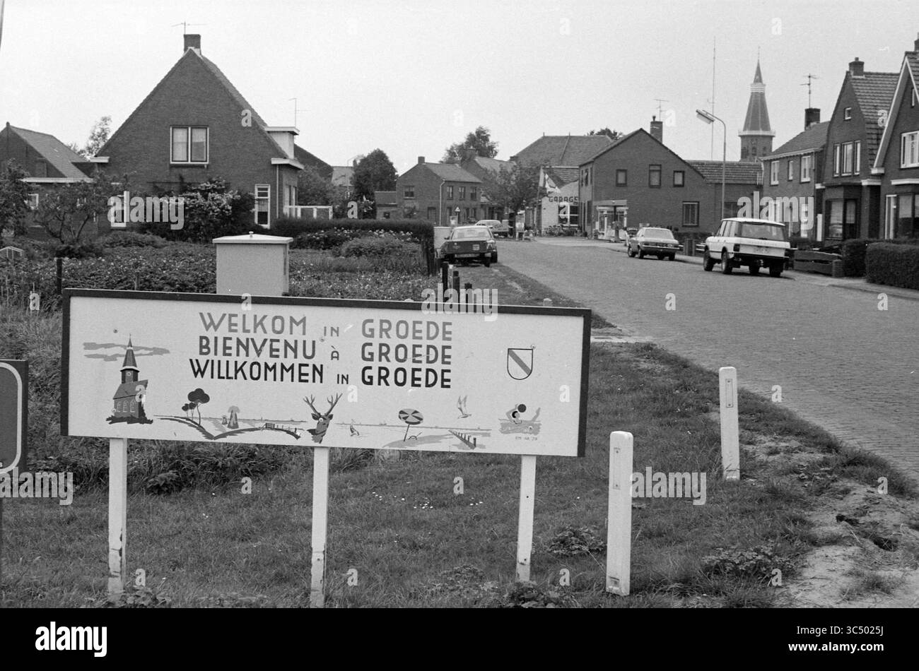 Rep. Groede, Zélande, villages et villes, Groede, 11-07-1984 Whizgle News, Dutch Desk, pays-Bas, 1950-2000 Un panneau de bienvenue à l'entrée d'un village pittoresque, affichant des salutations en plusieurs langues, flanqué de maisons simples et un aperçu d'un clocher d'église en arrière-plan le long d'une rue calme. Banque D'Images