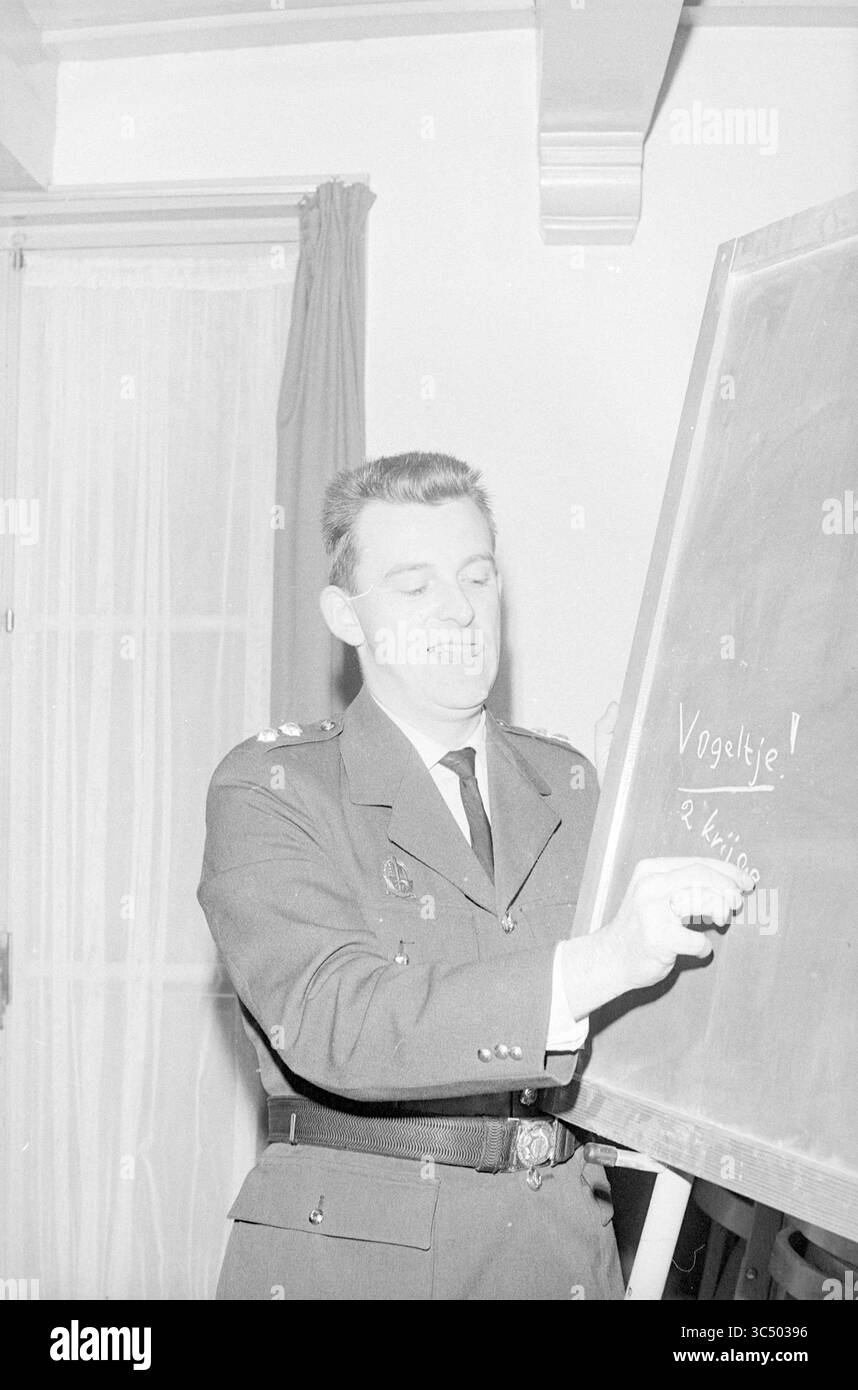 Portrait d'un officier de police Whizgle News, Dutch Desk, pays-Bas, 1950-2000 Un homme en tenue militaire écrit avec enthousiasme sur un tableau noir, s'engageant à enseigner ou expliquer quelque chose à son public. Le tableau comporte le mot 'Vogeltje !' et une note ou un nom d'accompagnement. Banque D'Images