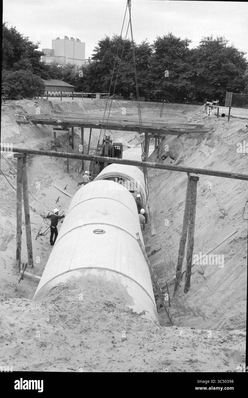 Construction d'un nouvel égout principal Whizgle News, Dutch Desk, pays-Bas, 1950-2000 Sur Un chantier de construction, des travailleurs sont positionnés près d'un grand tuyau cylindrique qui est descendu dans une tranchée. Des supports en bois encadrent l’excavation, tandis que la machinerie est visible en arrière-plan, soulignant l’atmosphère industrielle du projet. Banque D'Images