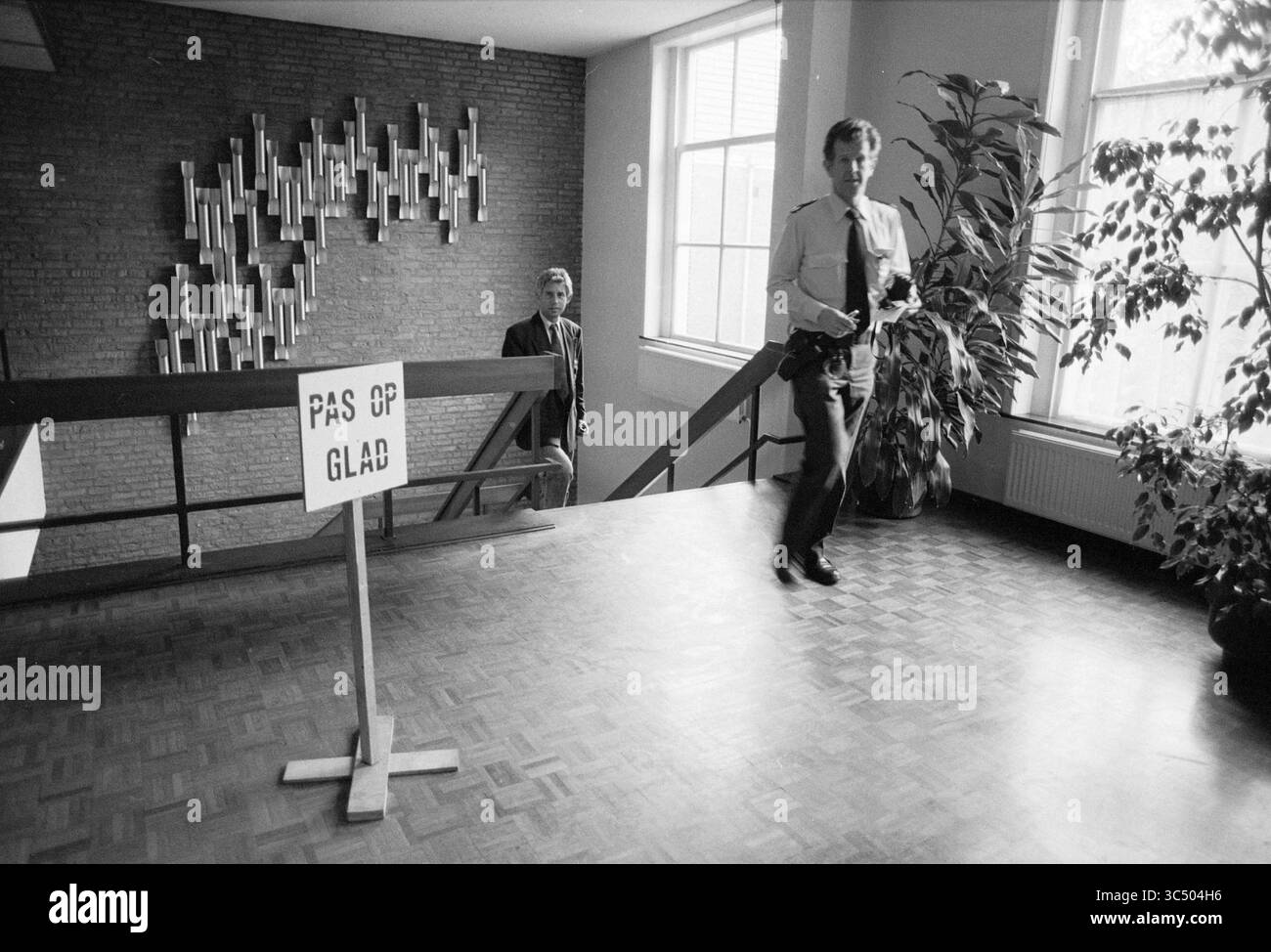 Smooth public Parquet's Office, police, 25-10-1989 Whizgle News, Dutch Desk, pays-Bas, 1950-2000 deux hommes sont vus dans un environnement intérieur spacieux. Un homme, vêtu d'uniforme, descend un escalier tandis que l'autre se tient au-dessus, observant. Un panneau indiquant « pas OP GLAD » est bien en vue au niveau inférieur, accompagné de grandes fenêtres et de verdure qui rehaussent l'atmosphère du cadre. Banque D'Images